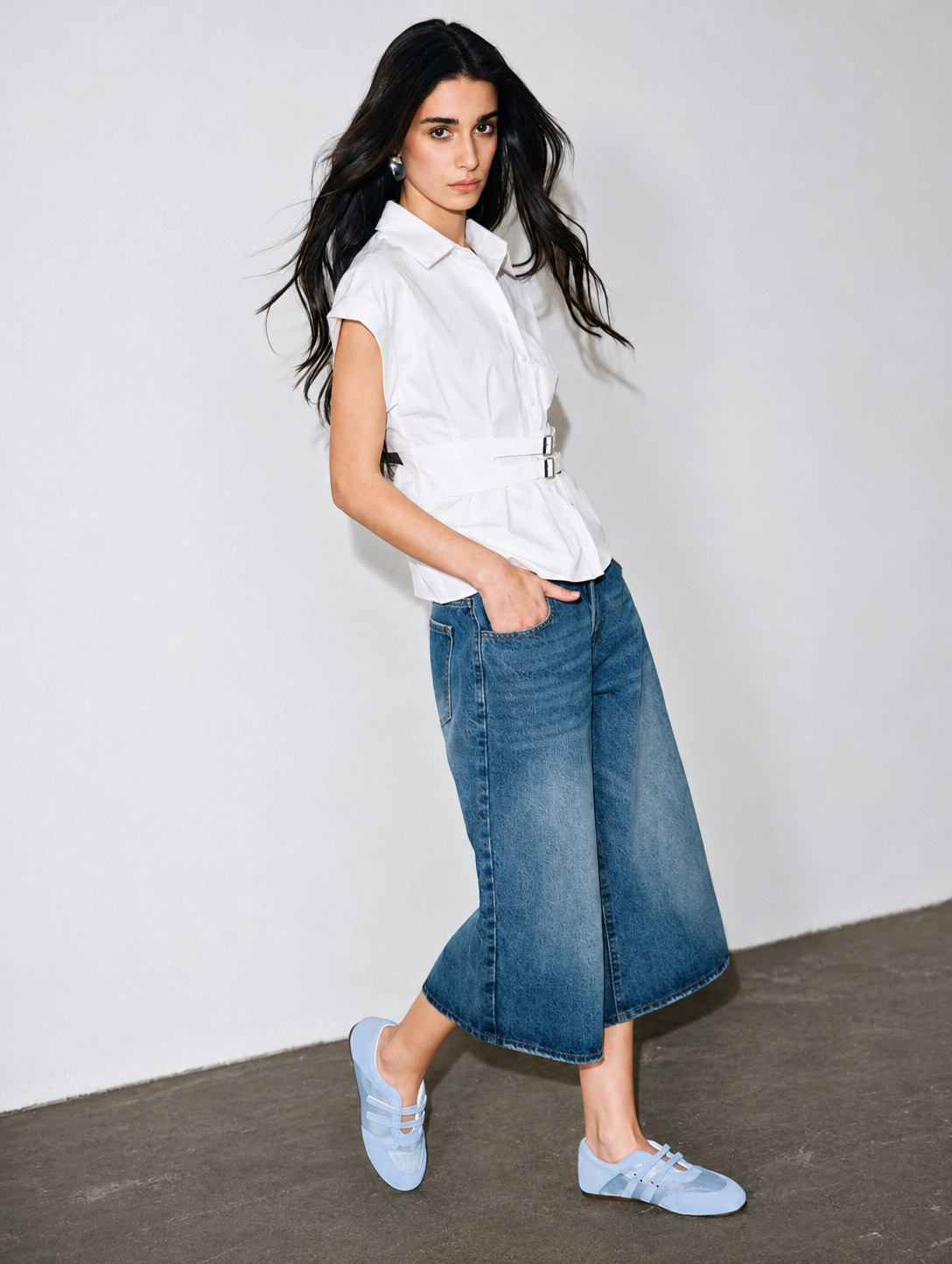 Mulher de cabelos longos e escuros, com express&atilde;o s&eacute;ria, vestindo camisa branca com cinto e cal&ccedil;a jeans culotte, t&ecirc;nis azuis claros, posando com uma m&atilde;o no bolso contra uma parede branca e ch&atilde;o cinza