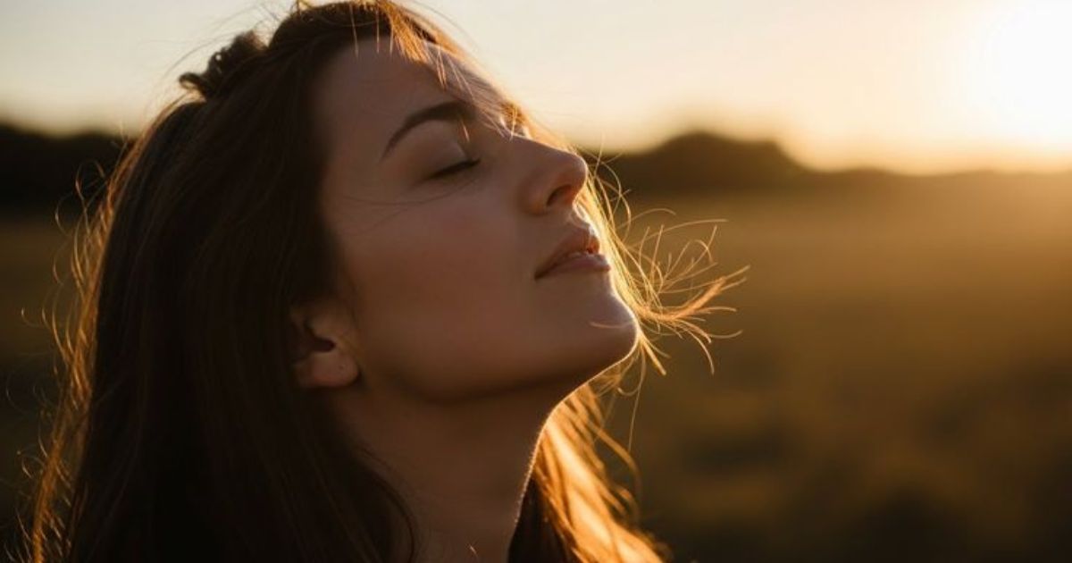 Mulher jovem de cabelos castanhos, olhos fechados e rosto erguido, com o sol do entardecer iluminando seu perfil e alguns fios de cabelo esvoa&ccedil;antes, em um campo com fundo desfocado