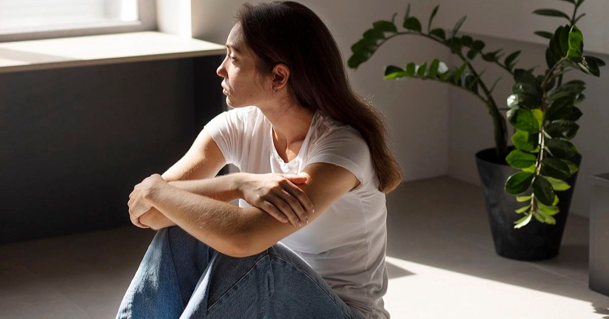 Mulher jovem de cabelos castanhos, camiseta branca e jeans, sentada no ch&atilde;o com os bra&ccedil;os cruzados, olhando pensativa para a janela. A luz do sol entra pela janela, iluminando parte do rosto e do corpo dela. Ao fundo, uma planta verde em vaso preto. O ambiente &eacute; claro e minimalista