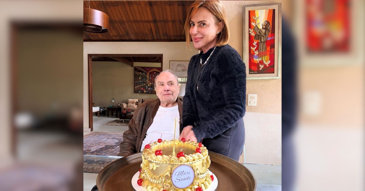 Uma mulher de cabelos curtos e ruivos, com blusa preta e colar de p&eacute;rolas, sorri ao lado de um homem sentado, de jaqueta marrom e camiseta branca. Eles est&atilde;o atr&aacute;s de um bolo dourado com cerejas e uma placa Mari Saade, em um ambiente interno com quadros e m&oacute;veis.