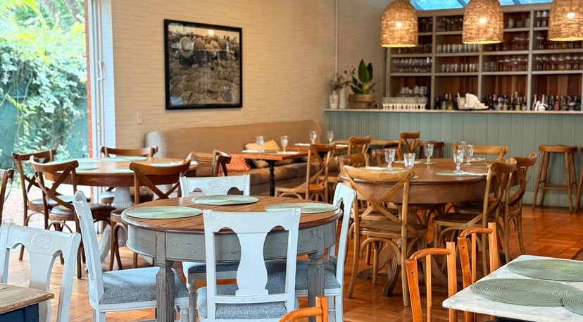 Interior de restaurante com mesas redondas de madeira e cadeiras variadas, algumas brancas e outras de madeira. H&aacute; um balc&atilde;o de bar ao fundo com prateleiras cheias de copos e garrafas, e lumin&aacute;rias de palha penduradas. Uma janela grande revela vegeta&ccedil;&atilde;o externa, e um quadro escuro decora a parede bege