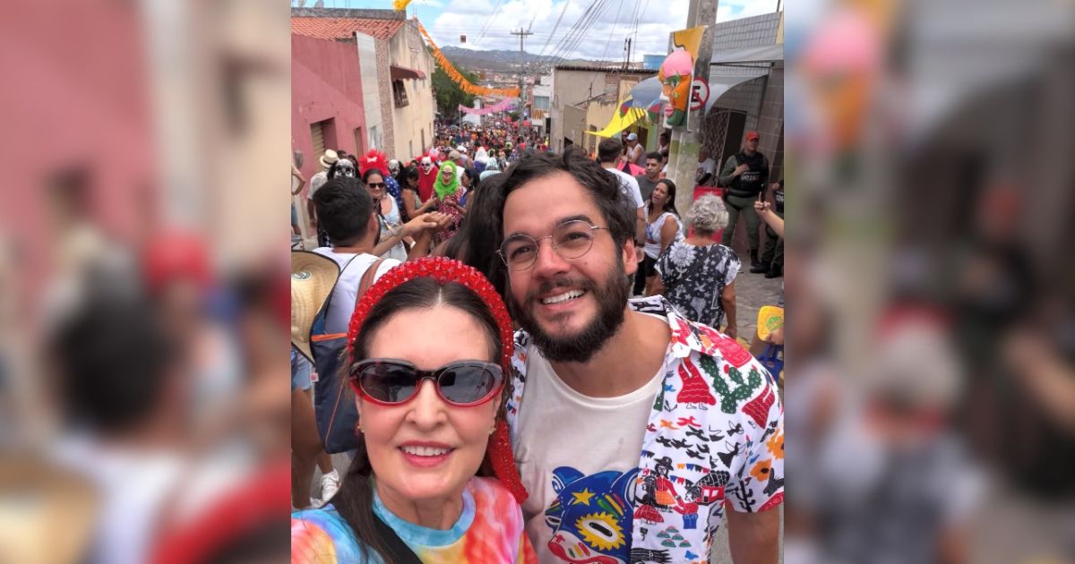 Mulher de &oacute;culos escuros e tiara vermelha sorri ao lado de homem barbudo de &oacute;culos, ambos em meio a uma multid&atilde;o em rua decorada para festa popular, com casas coloridas ao fundo