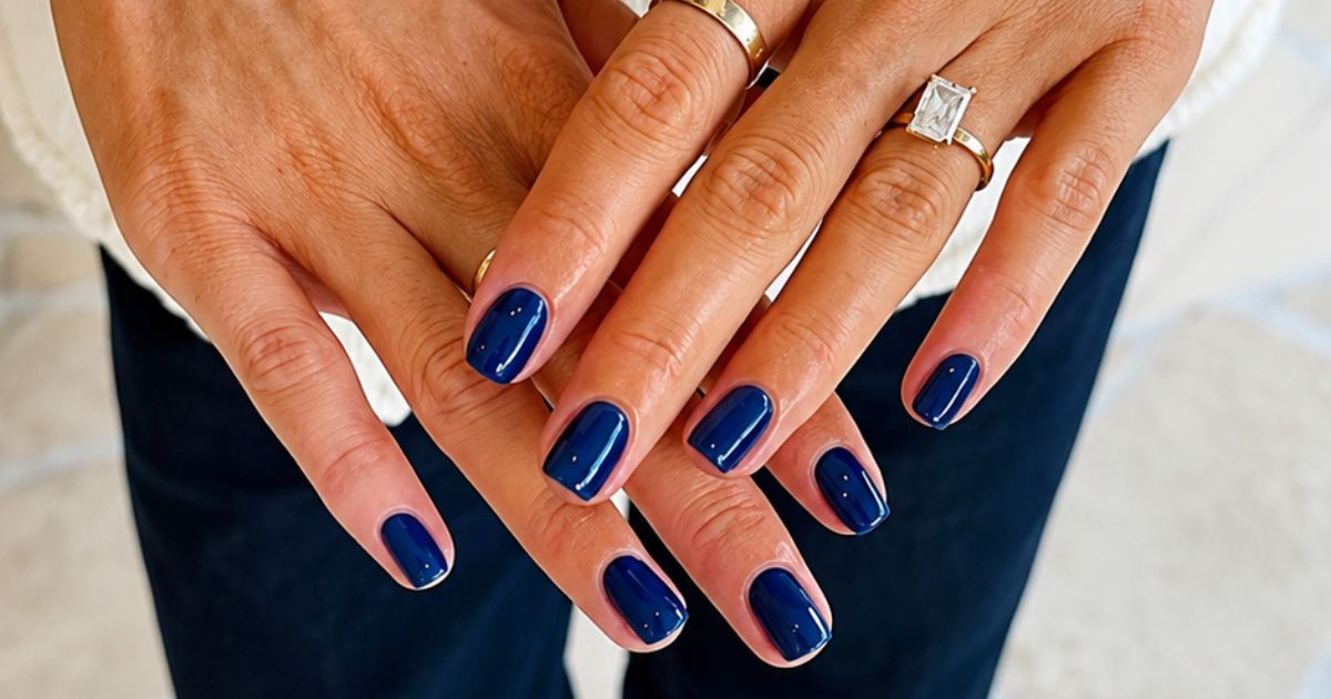 Duas m&atilde;os femininas com unhas curtas pintadas de azul marinho brilhante, uma com anel dourado liso e outra com anel dourado com pedra retangular transparente, sobre roupa escura e blusa clara