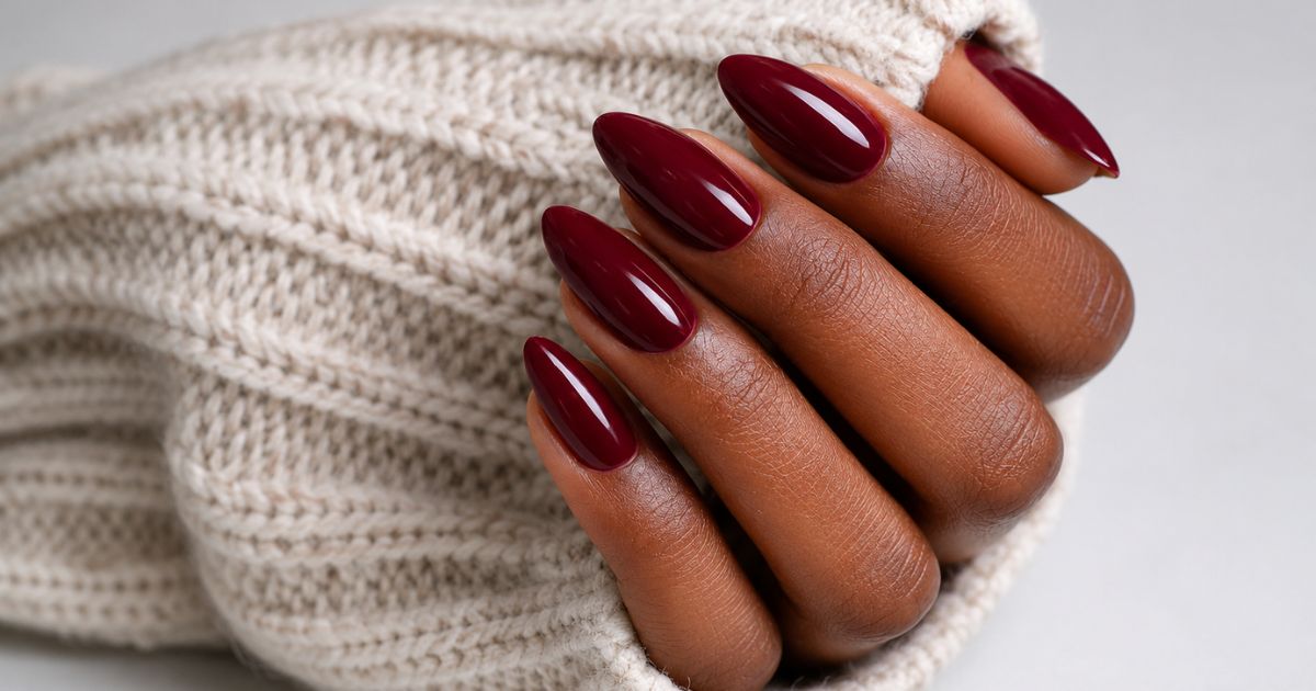M&atilde;o de pele escura com unhas longas e ovais pintadas de esmalte vinho brilhante, segurando um tecido de tric&ocirc; bege claro