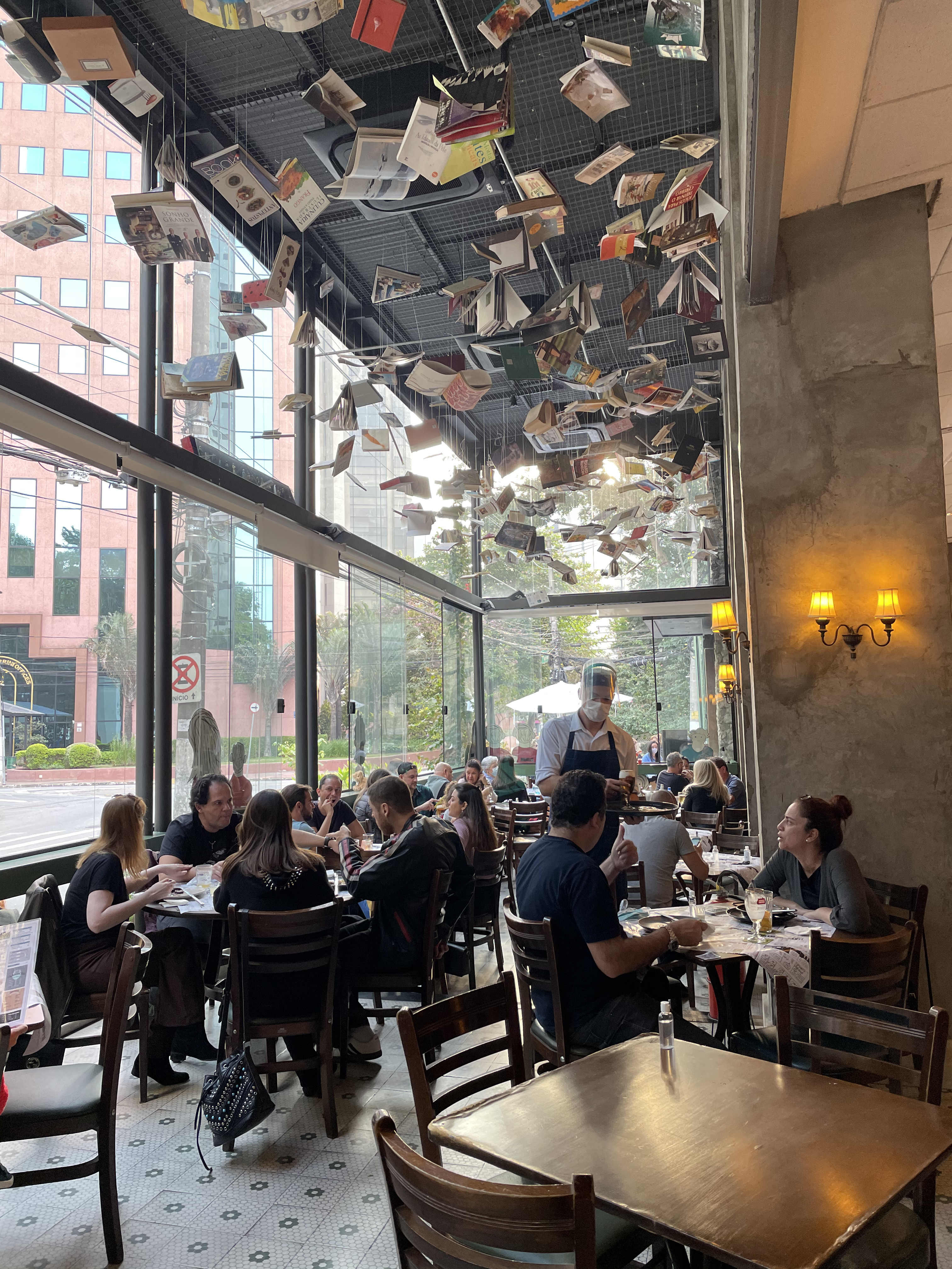 Interior de um restaurante movimentado com teto decorado por livros abertos suspensos. Clientes sentados em mesas de madeira, conversando e comendo, enquanto um gar&ccedil;om de m&aacute;scara facial serve. Grandes janelas de vidro mostram pr&eacute;dios e &aacute;rvores do lado de fora