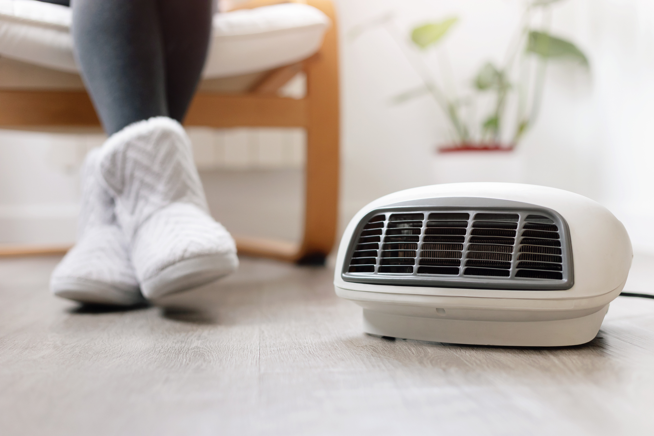 Pés de uma pessoa com meias pantufas brancas e cinzas, sentada em uma poltrona de madeira clara, próximos a um aquecedor elétrico branco no chão de madeira, com uma planta verde ao fundo