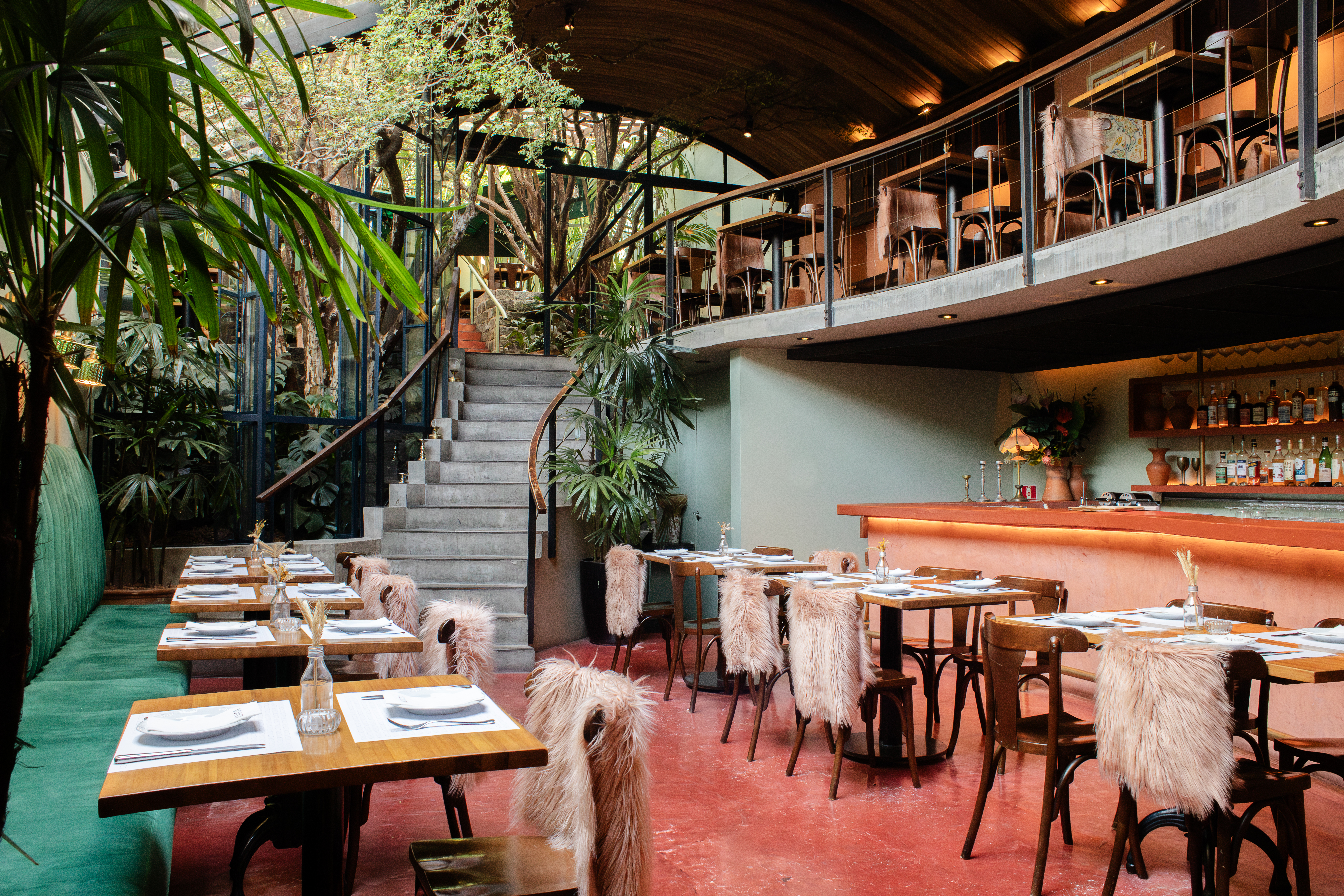 Restaurante com dois andares, mesas de madeira e cadeiras com capas de pele rosa, piso vermelho, bar laranja e plantas verdes. Uma escada de concreto leva ao andar superior, com mais mesas e um teto de madeira curvado. O ambiente &eacute; iluminado e aconchegante, com muitas plantas e &aacute;rvores vis&iacute;veis atrav&eacute;s de janelas de vidro