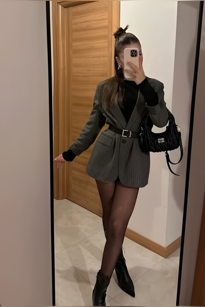Mulher jovem posando para selfie no espelho, usando blazer cinza risca de giz, cinto preto, meia-cal&ccedil;a escura, botas de bico fino e bolsa preta acolchoada. Ela tem cabelo castanho preso em coque alto e segura um celular claro que cobre seu rosto. Ao fundo, uma porta de madeira e parede branca