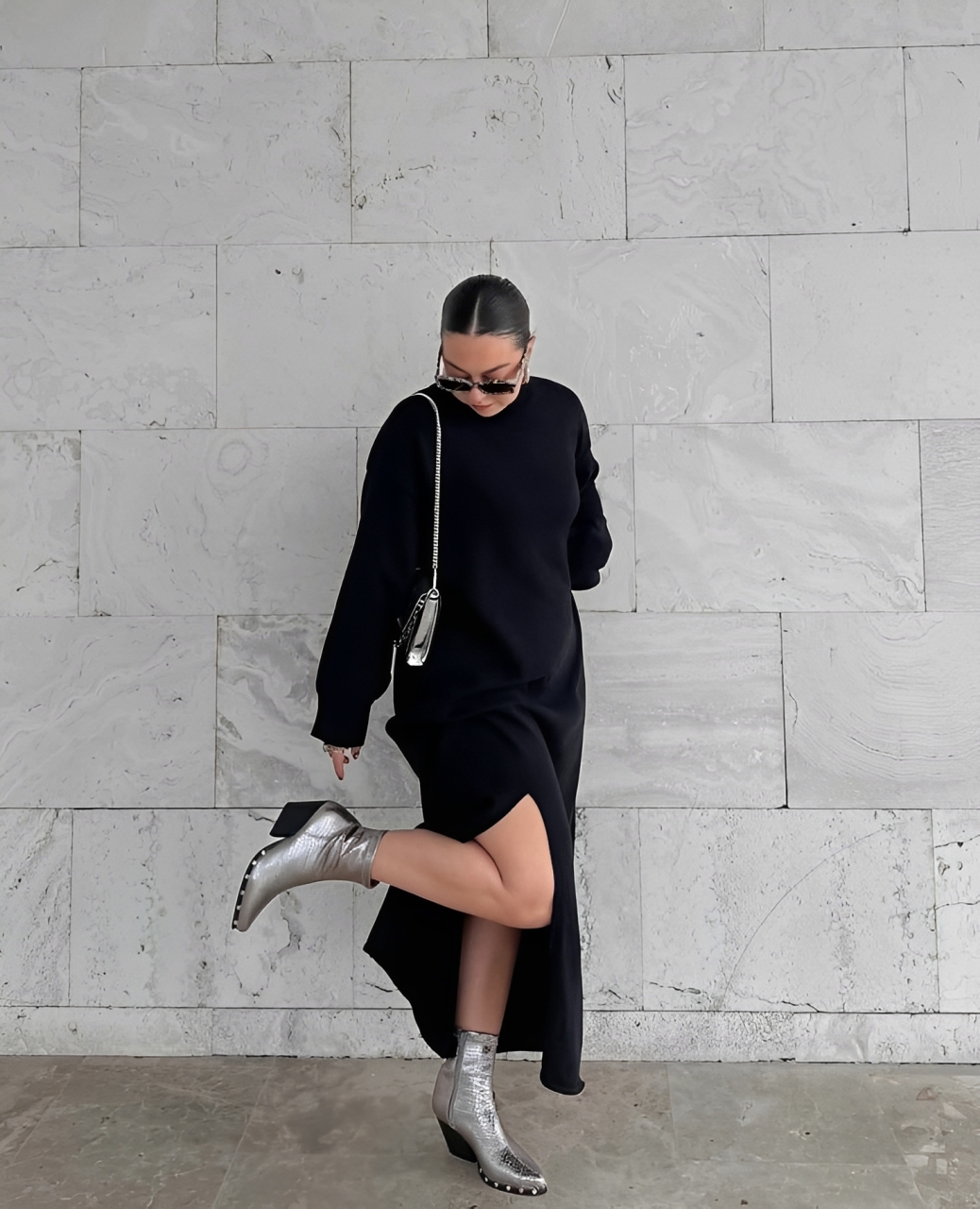 Mulher com coque, &oacute;culos escuros e vestido preto longo com fenda, posando com uma bota prateada levantada, em frente a uma parede de m&aacute;rmore branco
