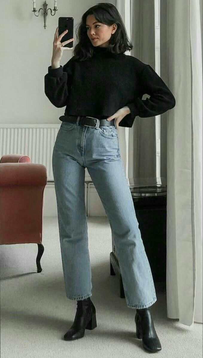 Mulher jovem com cabelo escuro na altura dos ombros, vestindo su&eacute;ter preto de gola alta, cal&ccedil;a jeans clara e botas pretas de salto, posa para selfie em um quarto claro com espelho e poltrona marrom