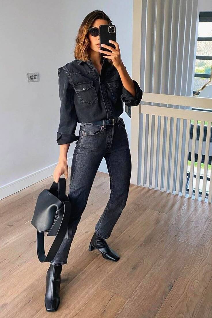 Mulher de cabelo castanho e &oacute;culos escuros tira selfie, vestindo camisa jeans escura, cal&ccedil;a jeans preta e botas pretas, segurando uma bolsa preta. Ela est&aacute; em um ambiente interno com piso de madeira e uma grade branca ao fundo