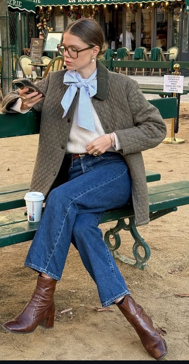 Mulher sentada em banco verde, usando &oacute;culos, jaqueta acolchoada marrom, su&eacute;ter creme, jeans e botas marrons. Ela segura um celular e tem um copo de caf&eacute; ao lado, com um caf&eacute; ao fundo