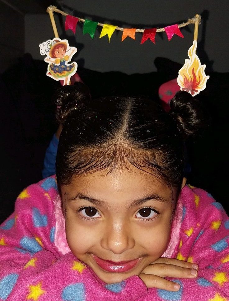 Menina sorridente com cabelo escuro preso em dois coques, usando um pijama rosa com estrelas amarelas e cora&ccedil;&otilde;es azuis. Ela tem decora&ccedil;&otilde;es de festa junina no cabelo: uma boneca caipira e uma fogueira, conectadas por uma cordinha com bandeirinhas coloridas. O rosto est&aacute; apoiado nas m&atilde;os, e ela olha diretamente para a c&acirc;mera