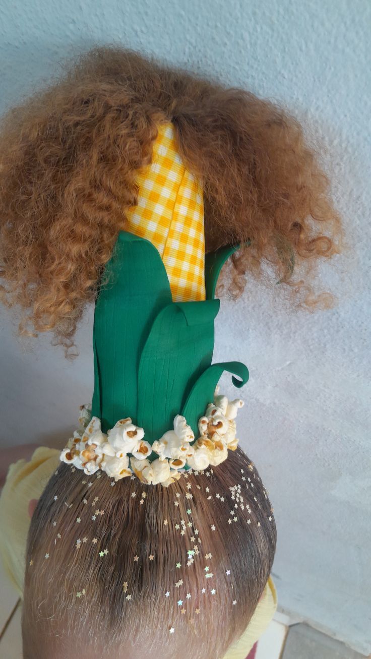 Crian&ccedil;a com cabelo castanho e brilhos de estrela, usando um chap&eacute;u de espiga de milho com cabelo cacheado ruivo e pipocas na base