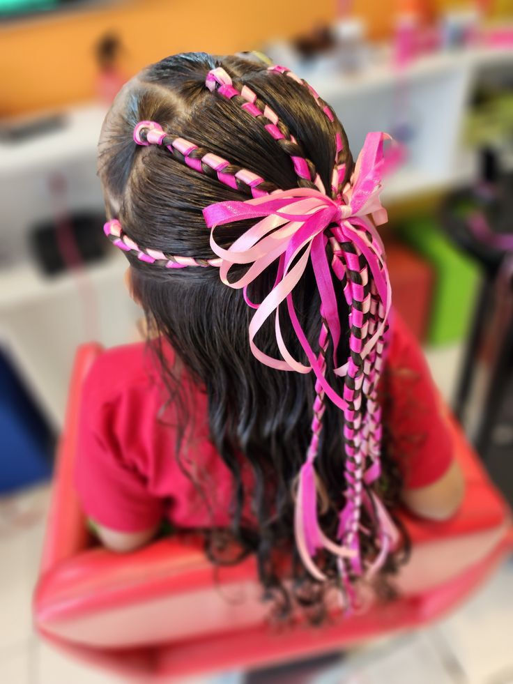 Crian&ccedil;a de costas com cabelo escuro, longo e ondulado, adornado com tran&ccedil;as finas e fitas rosa claro e pink, amarradas em um la&ccedil;o grande no topo da cabe&ccedil;a. Ela veste uma camiseta rosa e est&aacute; sentada em uma cadeira vermelha