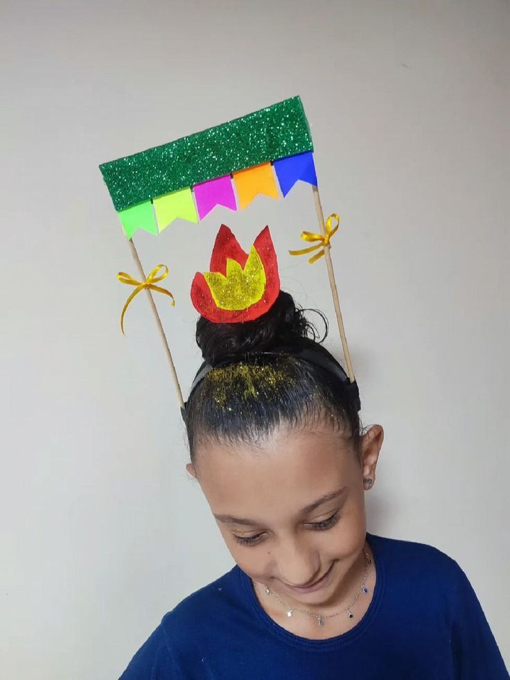 Menina com cabelo preso em coque, usando tiara com chap&eacute;u de festa junina: uma fogueira vermelha e amarela, e uma bandeirinha verde brilhante com outras coloridas penduradas. Ela sorri, olhando para baixo, e veste uma blusa azul com um colar prateado. O cabelo tem glitter dourado