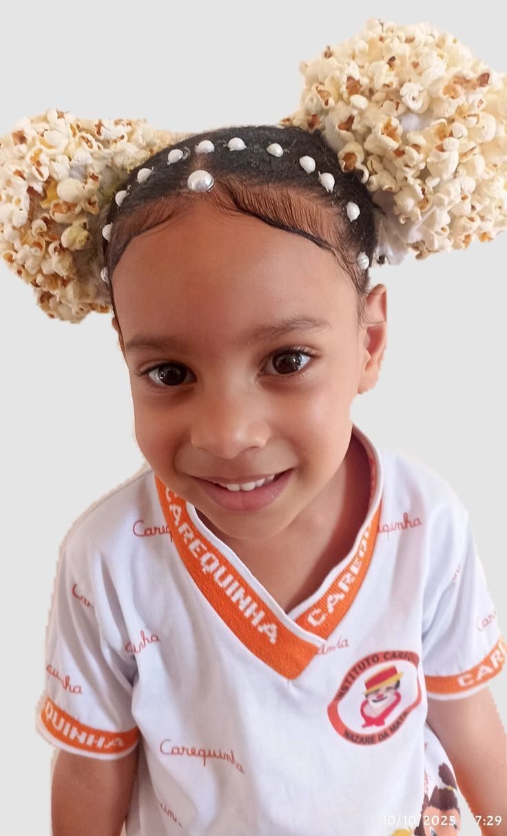 Crian&ccedil;a sorridente com cabelo preso em dois coques decorados com pipocas e pequenas p&eacute;rolas brancas, vestindo uma camiseta branca com gola e mangas laranjas, e o nome Carequinha estampado