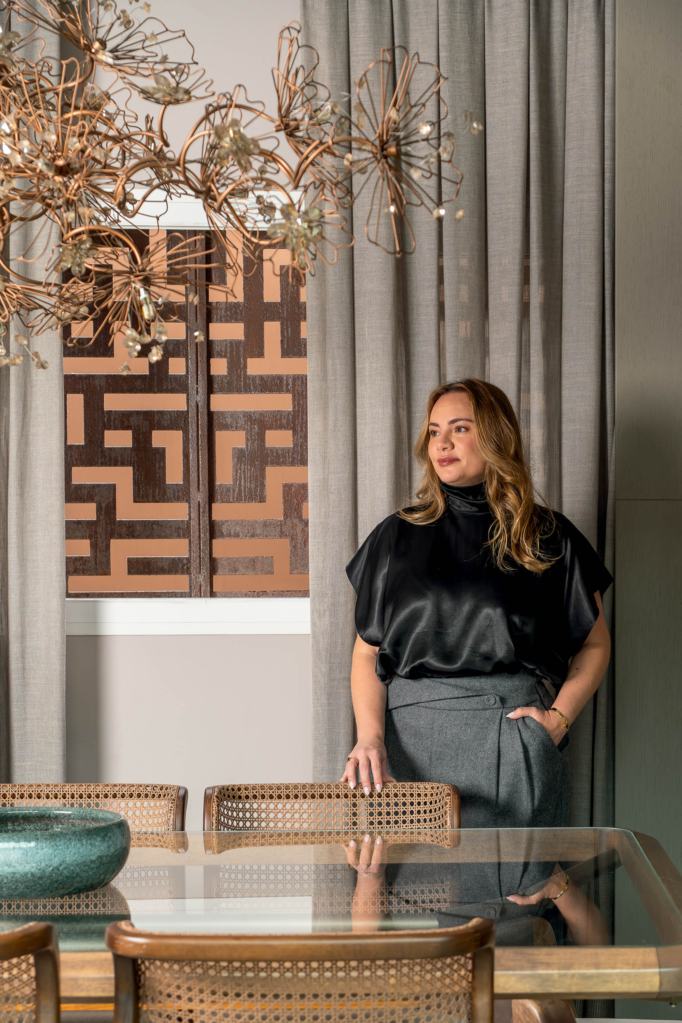 Mulher de cabelos castanhos claros, blusa preta de seda e cal&ccedil;a cinza, em p&eacute; ao lado de uma mesa de vidro com cadeiras de palha, olhando para a direita. Ao fundo, cortinas cinzas e uma janela com detalhes geom&eacute;tricos em tons de cobre. Um lustre de metal e cristais pende do teto