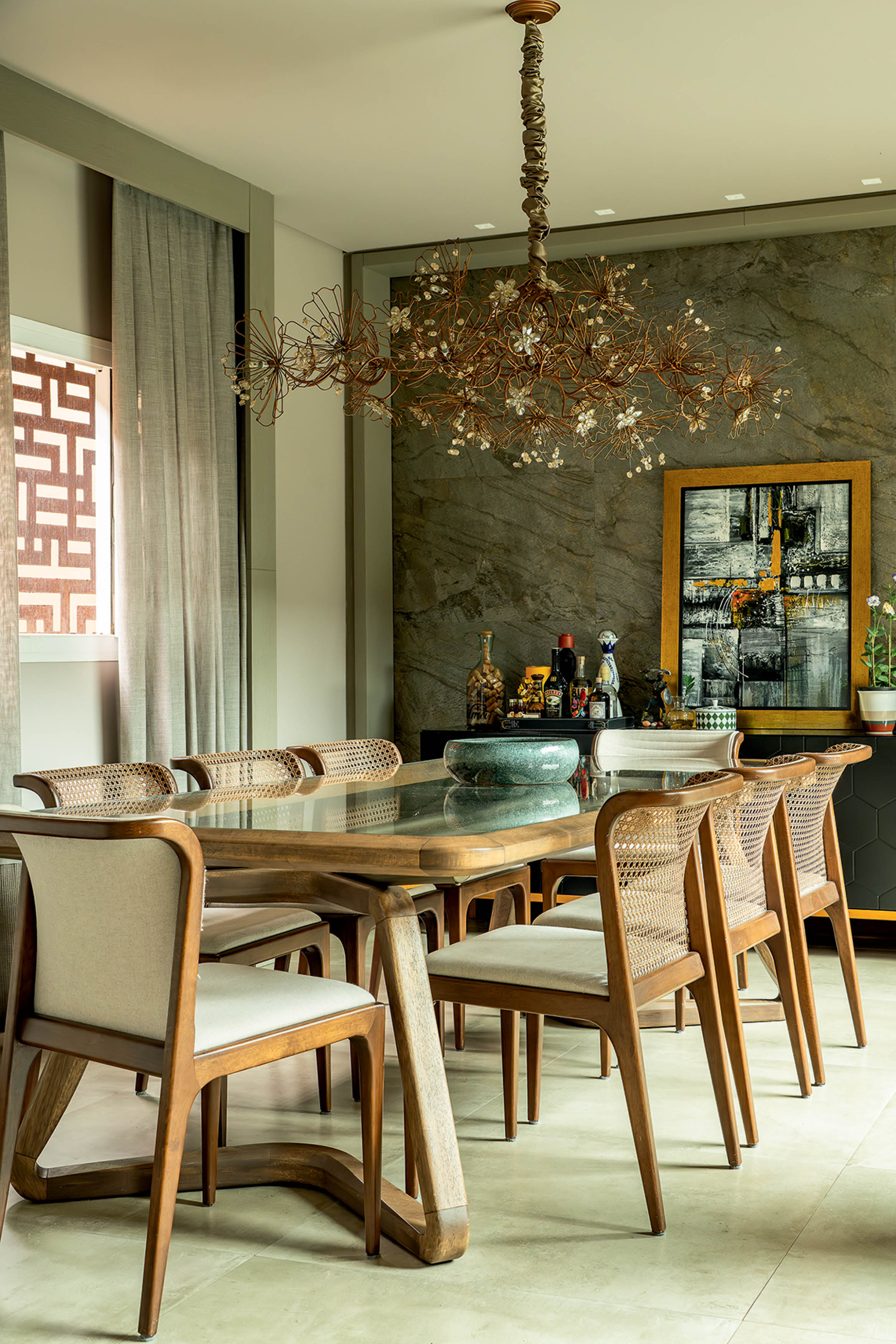 Sala de jantar com mesa de madeira e tampo de vidro, cercada por cadeiras de madeira com assentos estofados claros e encostos de palha. Um lustre de galhos e flores brancas pende sobre a mesa, que tem uma tigela verde. Ao fundo, uma parede de pedra escura exibe um quadro abstrato em tons de cinza e amarelo, e um aparador com garrafas e objetos decorativos. Cortinas cinzas e uma janela com treli&ccedil;a geom&eacute;trica completam o ambiente