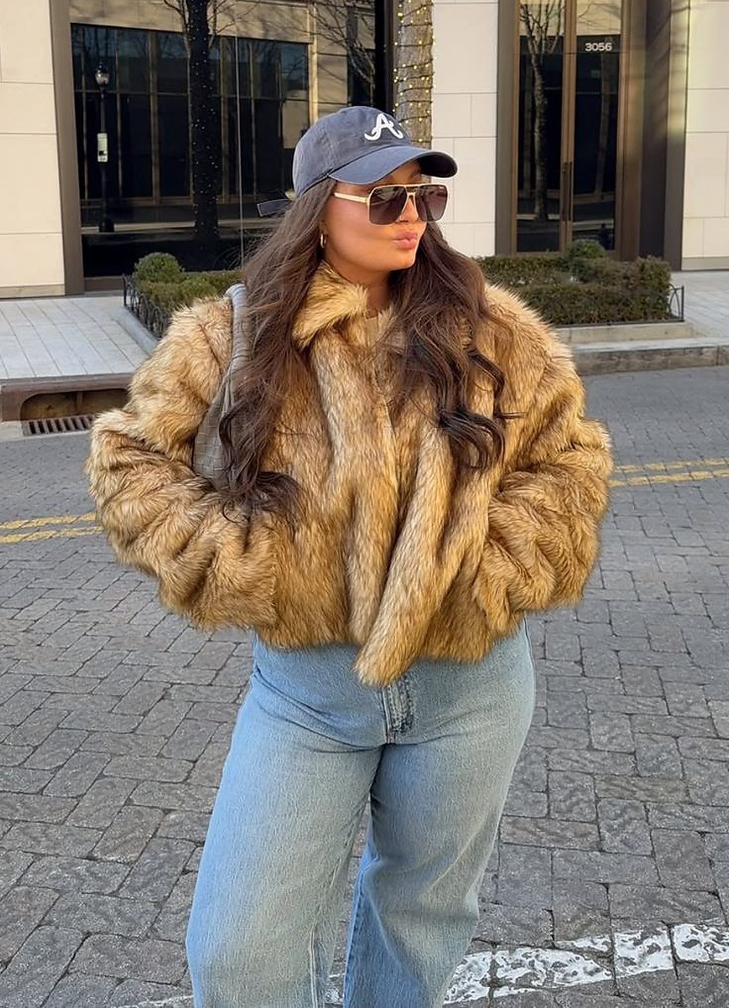 Mulher jovem, cabelos longos e castanhos, vestindo casaco de pele sint&eacute;tica marrom, cal&ccedil;a jeans clara, bon&eacute; azul com A branco e &oacute;culos de sol, posa com as m&atilde;os nos bolsos e boca de beijo em rua de paralelep&iacute;pedos, com pr&eacute;dios e uma &aacute;rvore iluminada ao fundo