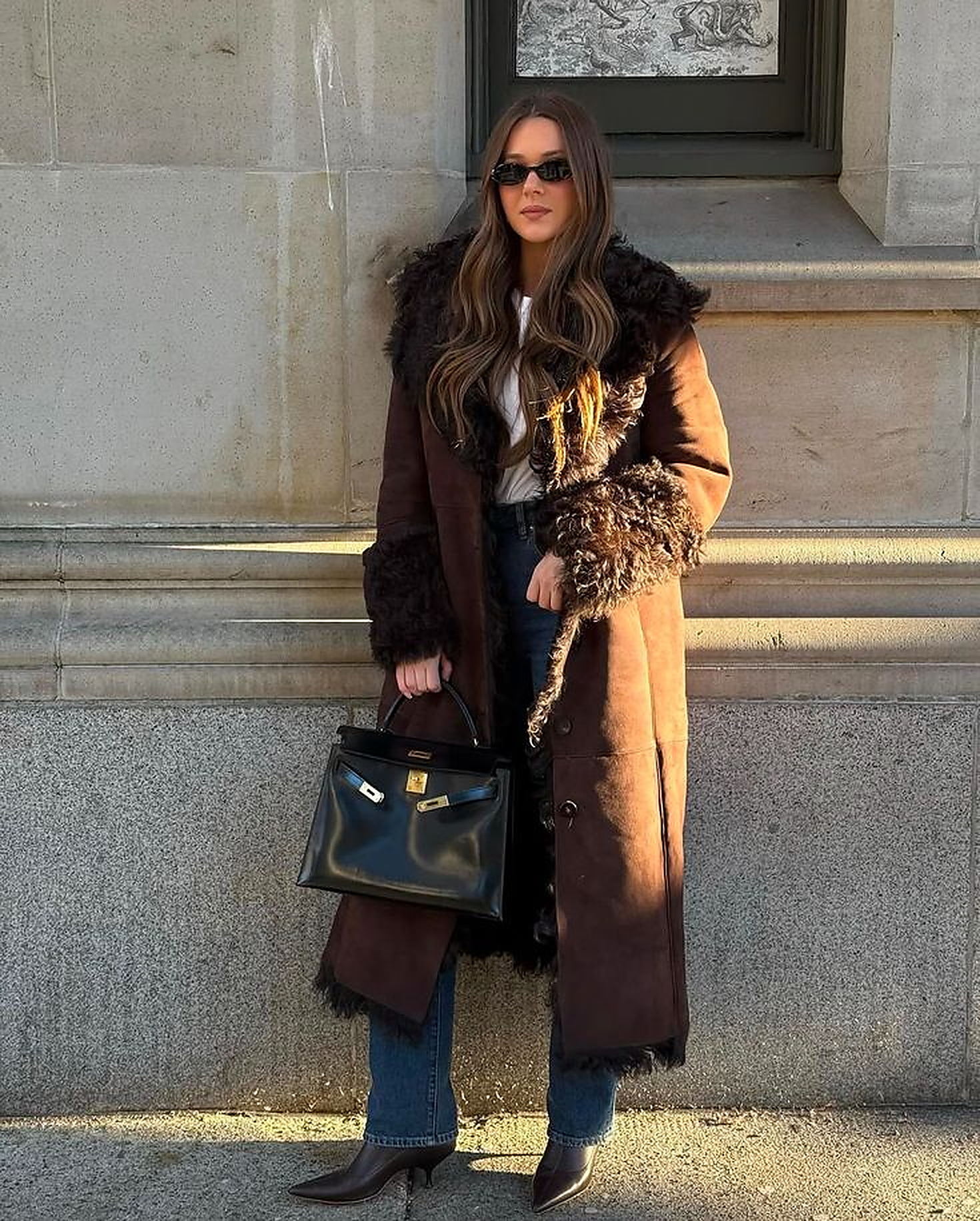 Mulher de cabelos castanhos longos, &oacute;culos escuros, casaco marrom de camur&ccedil;a com detalhes de pele escura, jeans e botas de salto baixo, segurando uma bolsa preta com fechos dourados, posando em frente a uma parede de pedra sob a luz do sol
