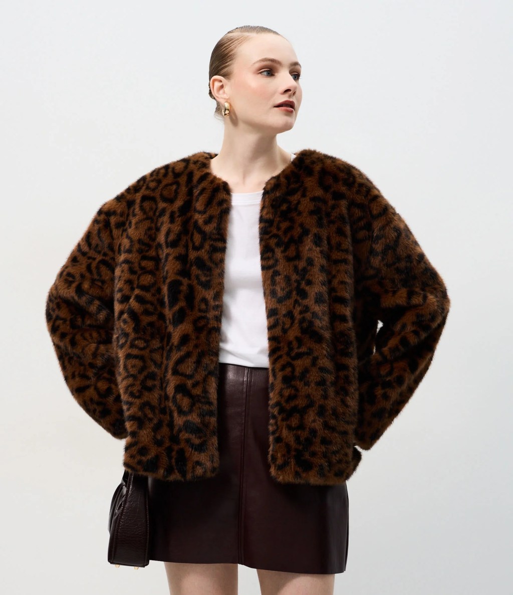 Mulher loira com coque elegante, usando casaco de pele sint&eacute;tica com estampa de leopardo, camiseta branca e saia de couro marrom, posa com uma bolsa pequena escura no bra&ccedil;o esquerdo