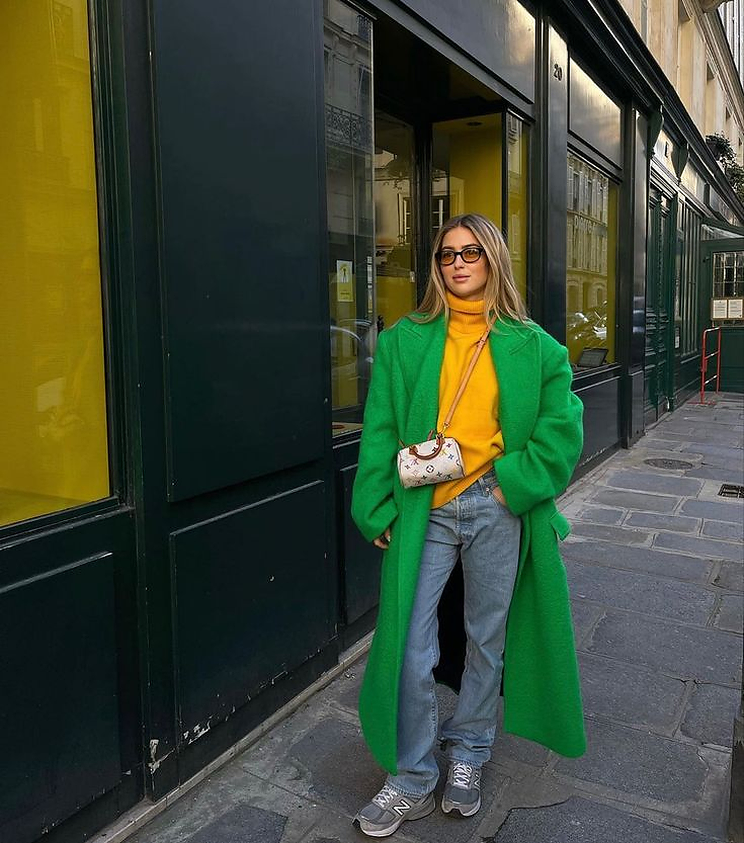 Mulher loira de &oacute;culos escuros, casaco verde-esmeralda longo, su&eacute;ter amarelo-mostarda, jeans claro e t&ecirc;nis cinza, posando em cal&ccedil;ada de paralelep&iacute;pedos com lojas de fachada escura e vitrines amarelas ao fundo