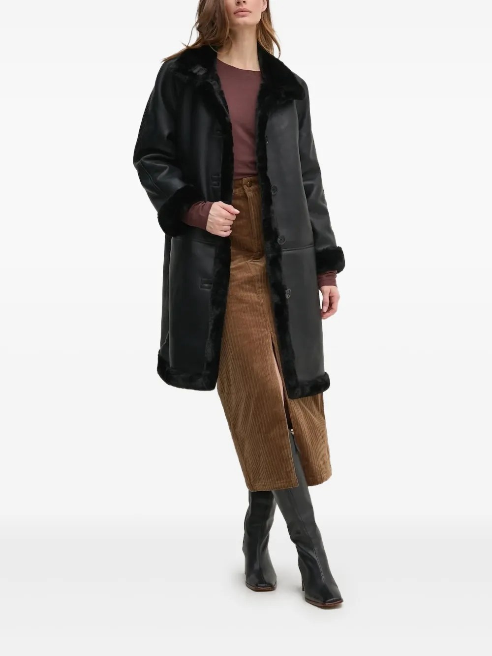 Mulher de cabelos castanhos, em p&eacute;, vestindo um casaco longo preto com detalhes de pele sint&eacute;tica preta, blusa marrom, saia midi de veludo cotel&ecirc; marrom com fenda frontal e botas pretas de cano alto