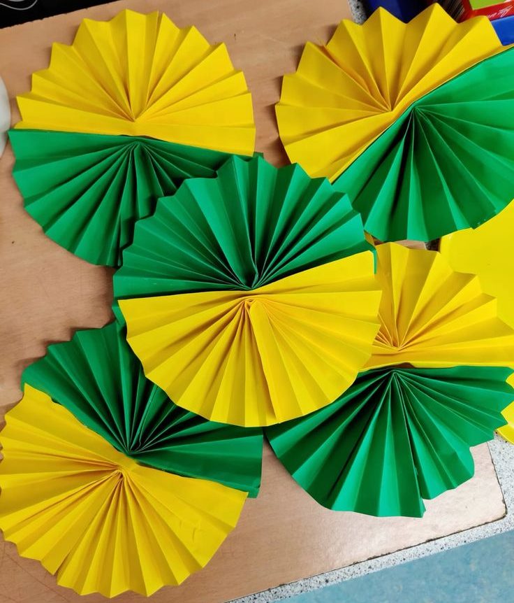 V&aacute;rios leques de papel artesanais, metade verde e metade amarelo, dispostos sobre uma superf&iacute;cie de madeira clara