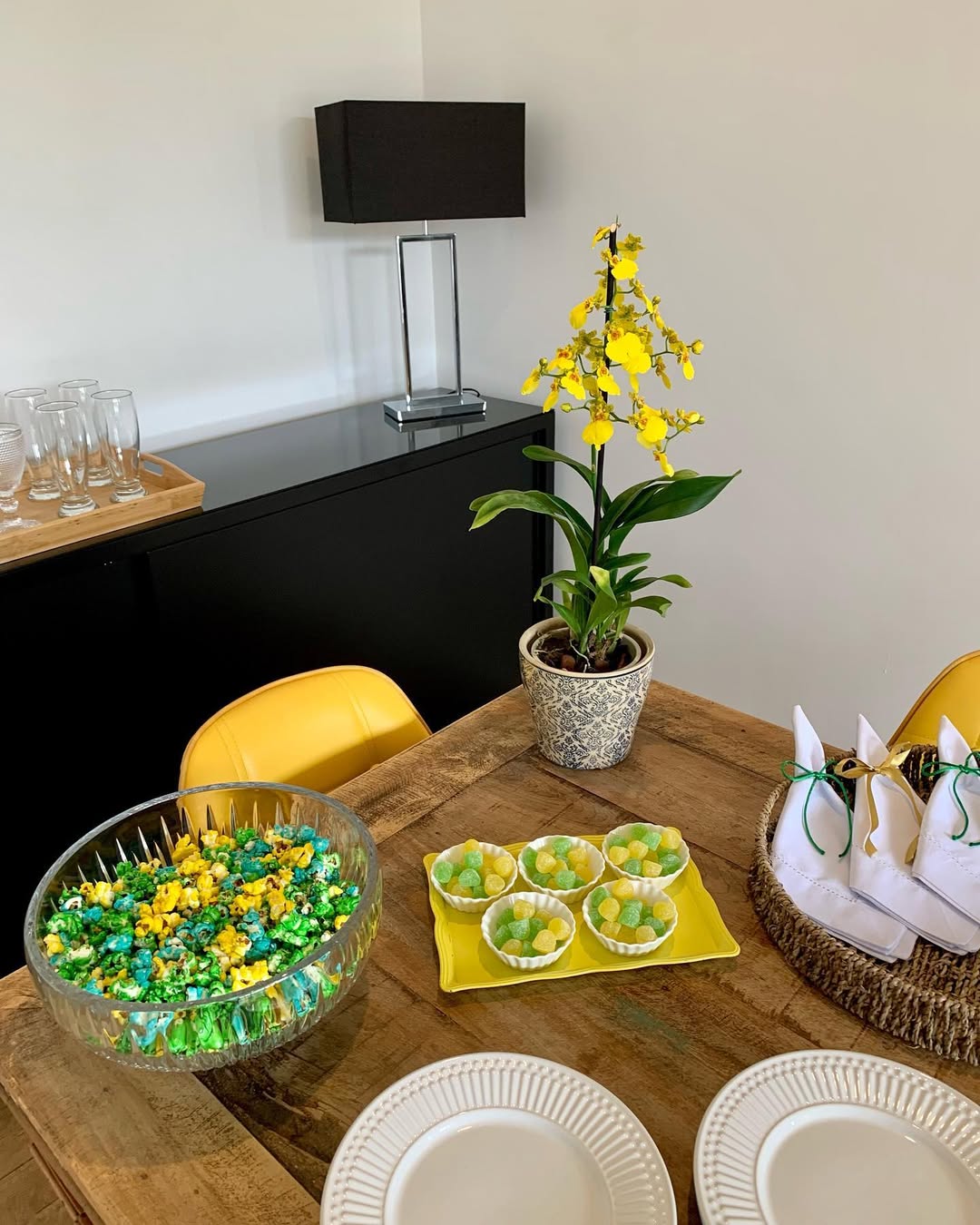 Mesa de madeira r&uacute;stica com tigela de pipoca colorida, doces amarelos e verdes em prato retangular, e guardanapos brancos em cesto. Ao fundo, orqu&iacute;dea amarela em vaso, abajur preto e copos em bandeja sobre m&oacute;vel escuro