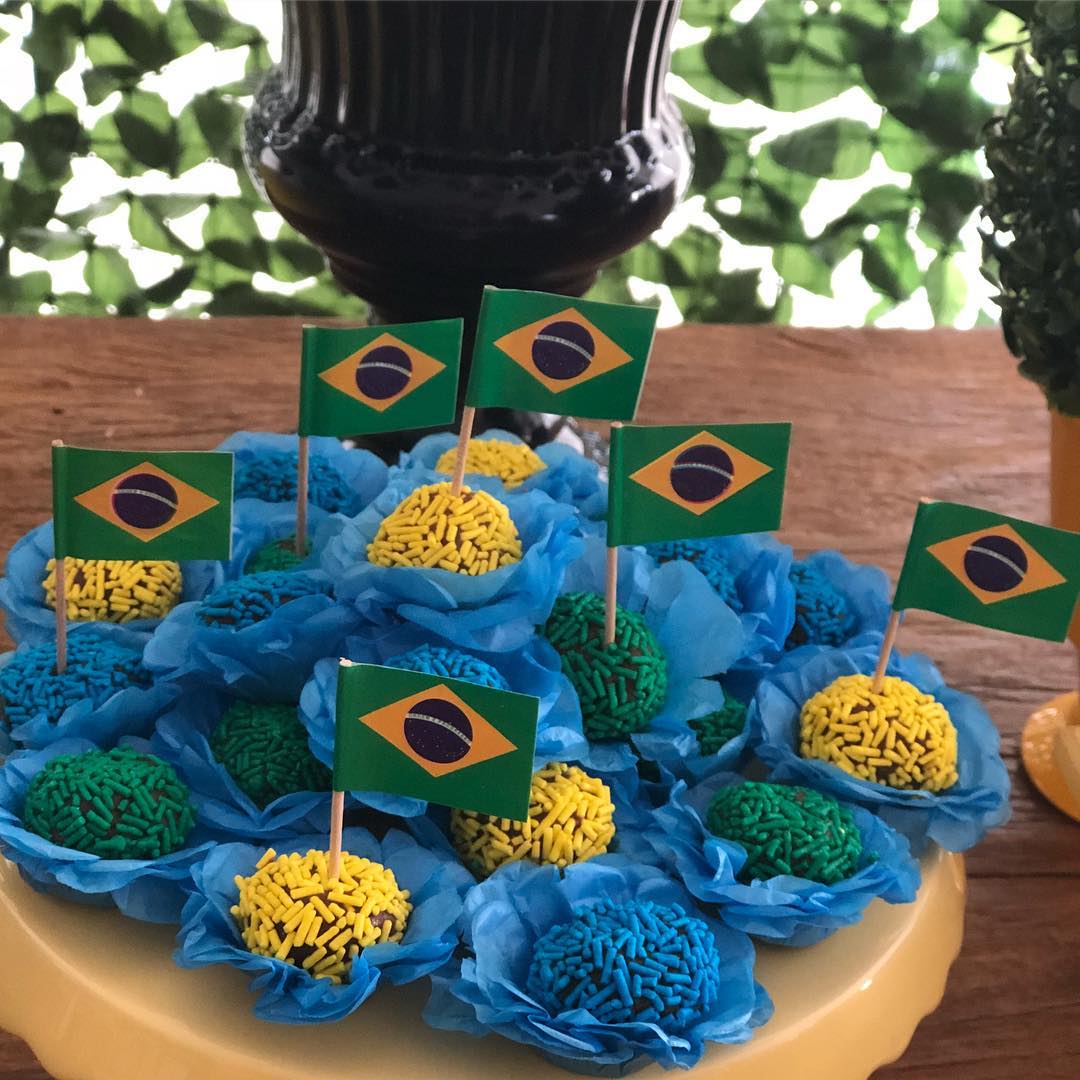 Docinhos de festa brasileiros, brigadeiros e beijinhos, com granulados amarelos, verdes e azuis, em forminhas azuis, decorados com pequenas bandeiras do Brasil. Est&atilde;o sobre um prato amarelo, com fundo de folhagem verde e um vaso preto