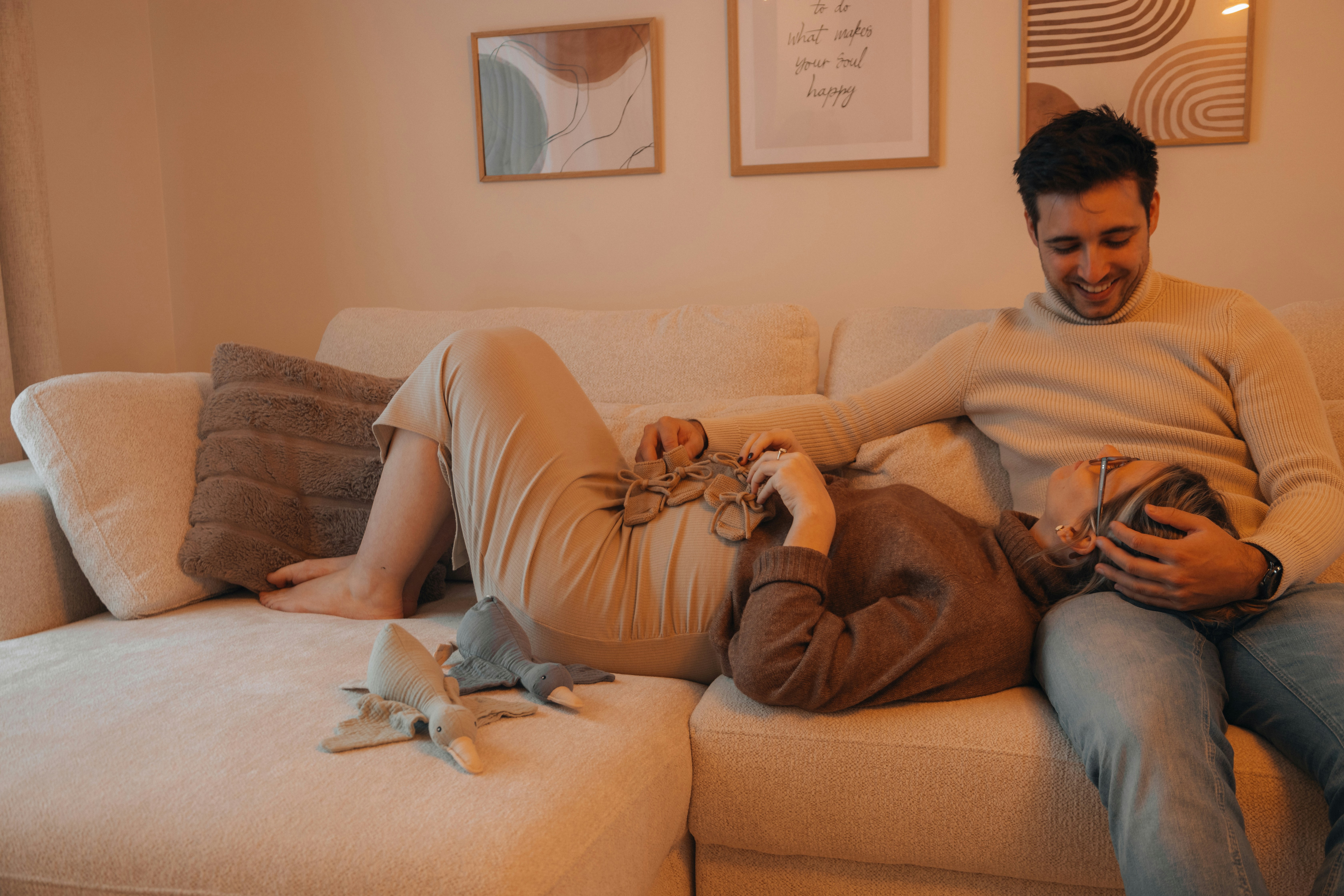 Casal relaxando no sof&aacute; bege: mulher gr&aacute;vida deitada com a cabe&ccedil;a no colo do homem, que sorri e a abra&ccedil;a. Ela segura sapatinhos de beb&ecirc; na barriga, enquanto dois patos de pel&uacute;cia est&atilde;o no sof&aacute;. Quadros abstratos decoram a parede.