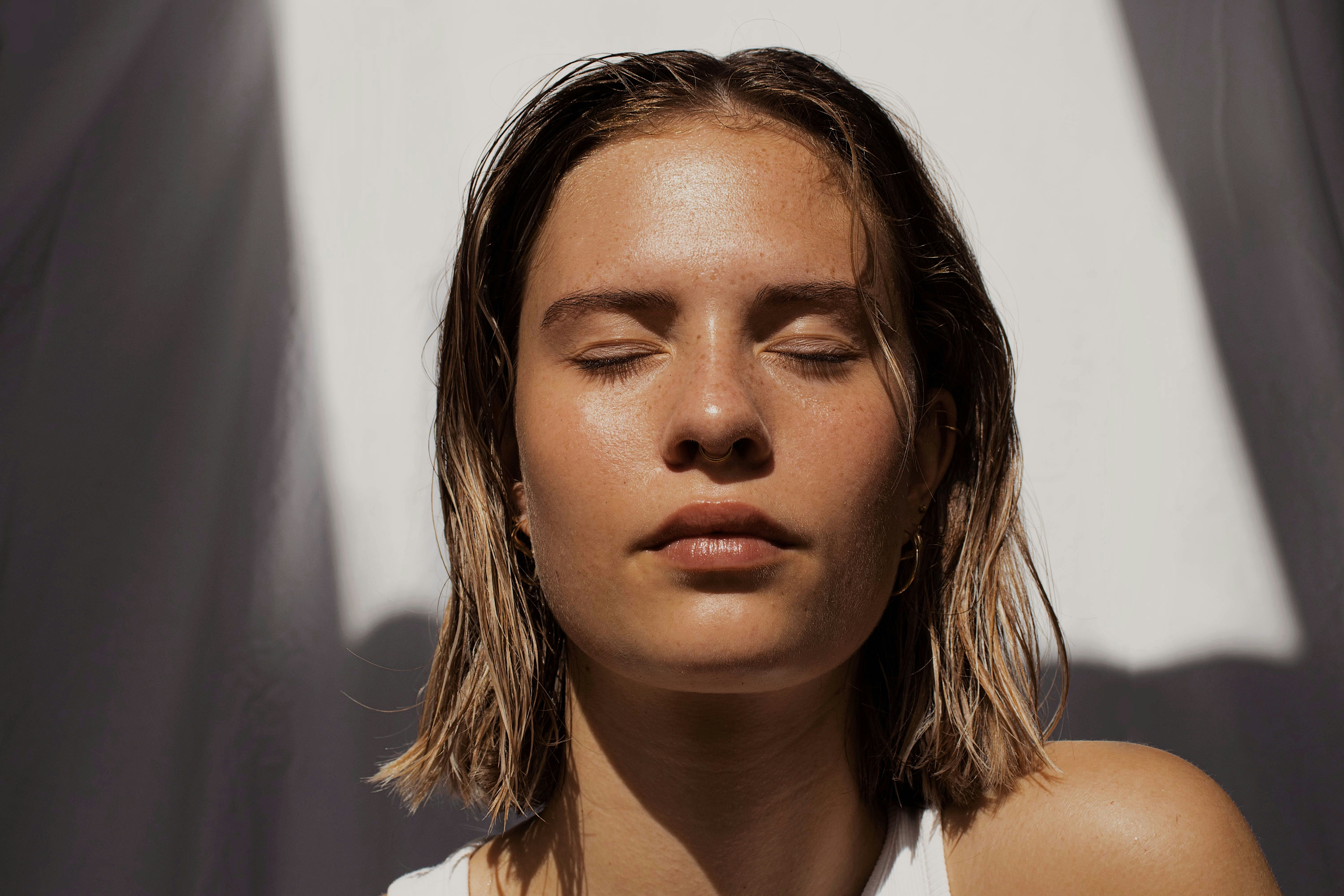 Mulher jovem com cabelo molhado e curto, olhos fechados, rosto com sardas e pele bronzeada, iluminada por um feixe de luz solar diagonal em um fundo branco e cinza
