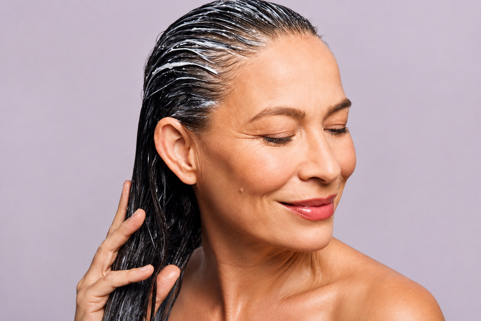 Mulher de meia-idade com cabelos escuros molhados e creme branco aplicado no couro cabeludo, sorrindo com olhos fechados, tocando os fios com a m&atilde;o direita, ombros nus, em fundo lil&aacute;s