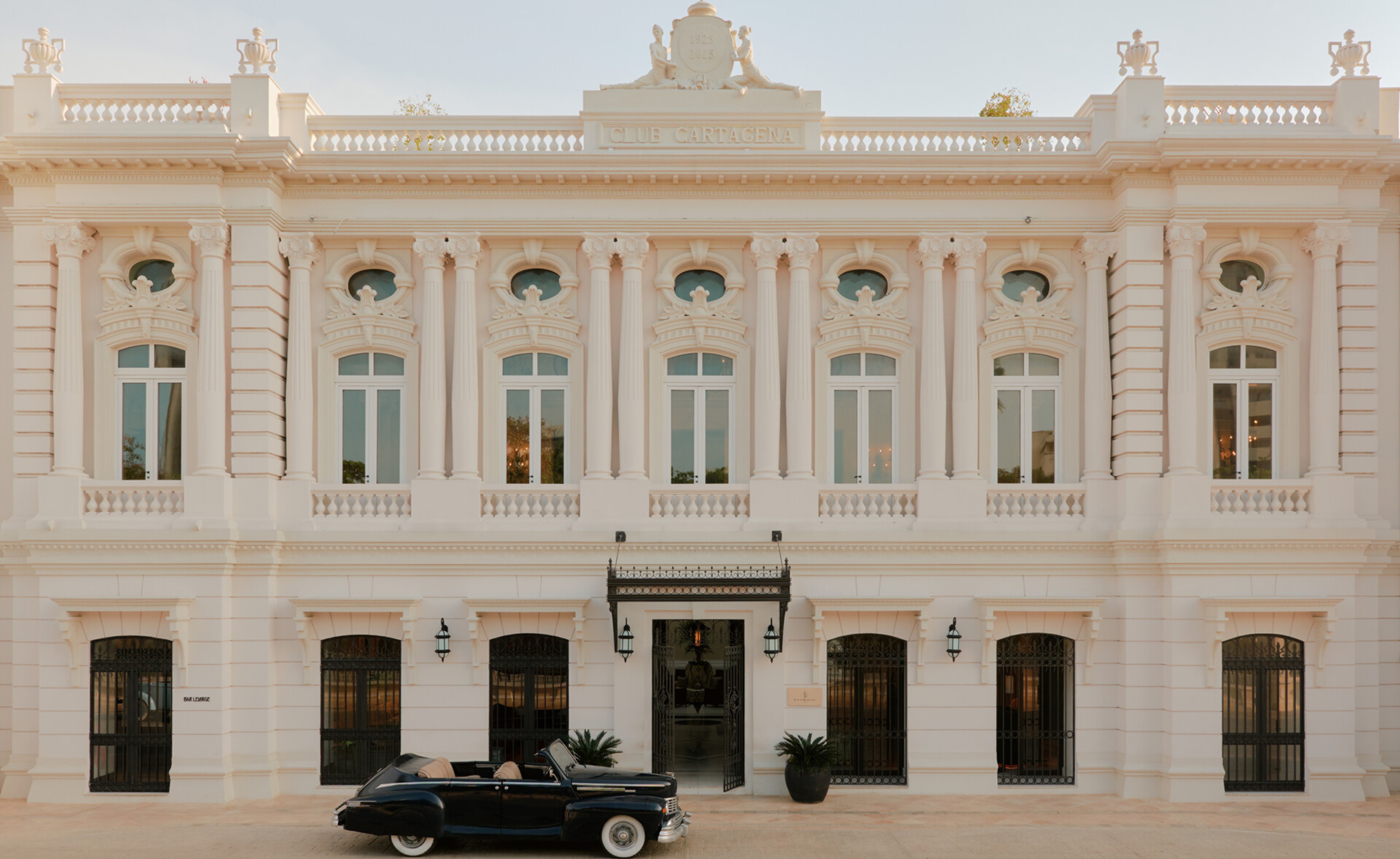 Fachada de um edif&iacute;cio neocl&aacute;ssico branco com CLUB CARTAGENA no topo, janelas altas e sacadas com balaustradas. Um carro convers&iacute;vel preto antigo est&aacute; estacionado em frente &agrave; entrada principal, que possui um toldo de ferro forjado.