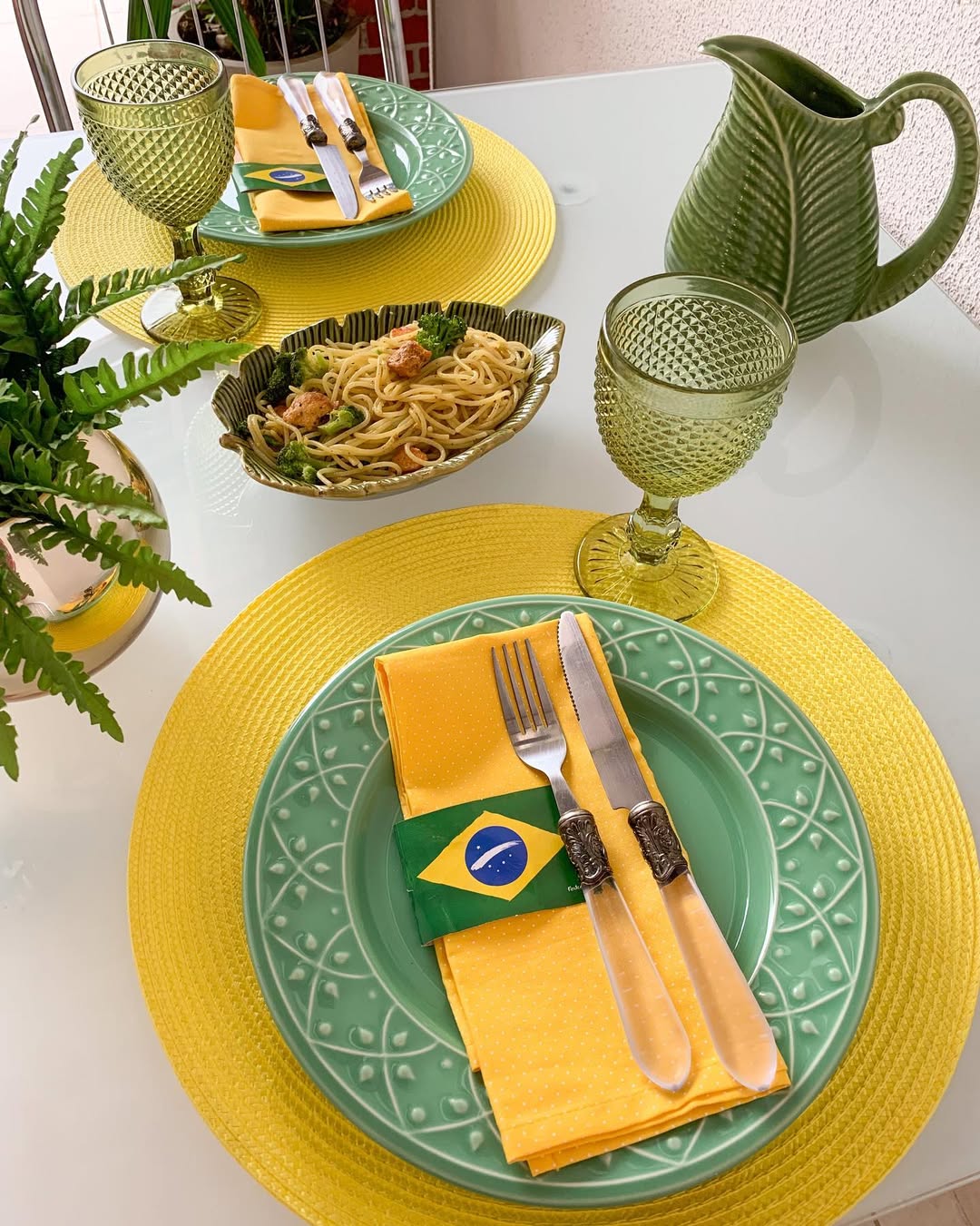 Mesa posta com dois lugares, pratos verdes sobre jogos americanos amarelos, guardanapos amarelos com an&eacute;is da bandeira do Brasil. H&aacute; talheres, ta&ccedil;as verdes, um jarro verde em formato de folha e uma travessa com macarr&atilde;o, br&oacute;colis e frango