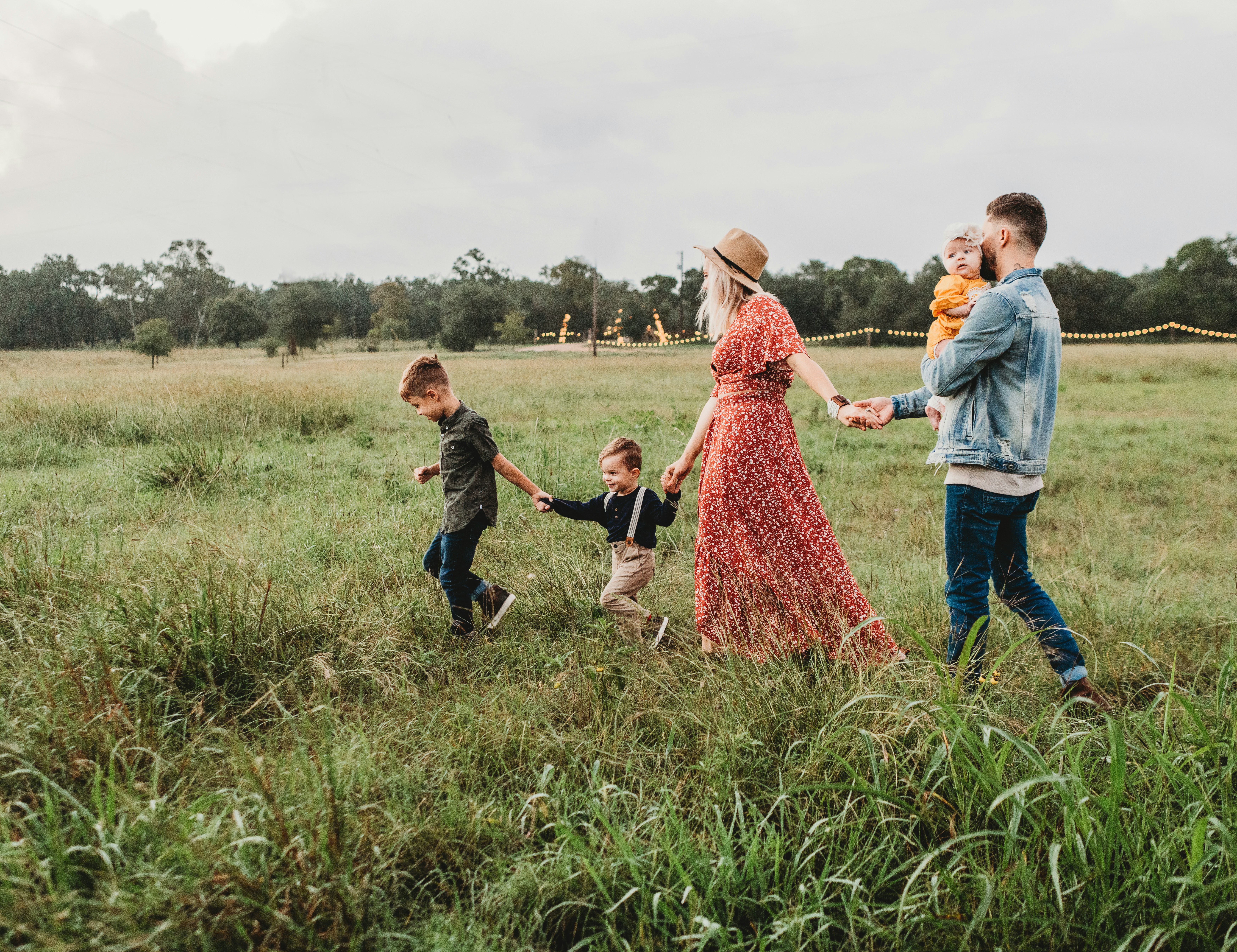 Fam&iacute;lia de cinco pessoas caminhando em um campo verde. A m&atilde;e, de vestido floral vermelho e chap&eacute;u, segura a m&atilde;o do filho mais novo, de camisa preta e suspens&oacute;rios. O filho do meio, de camisa verde escura, segura a m&atilde;o do irm&atilde;o mais novo. O pai, de jaqueta jeans, carrega um beb&ecirc; de roupa amarela.