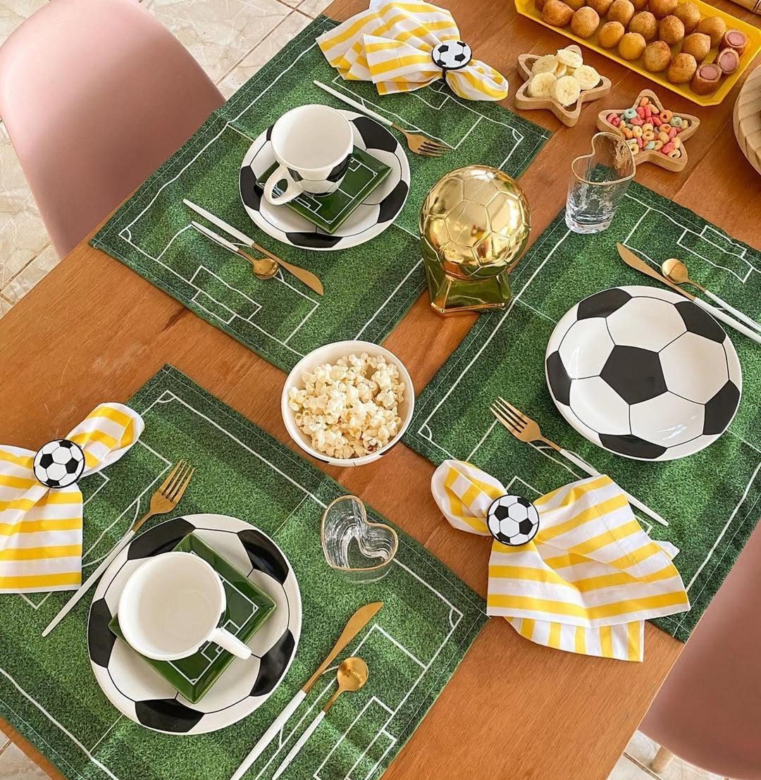 Mesa de madeira posta para festa de futebol com quatro lugares. Cada lugar tem um jogo americano verde com linhas de campo, prato de bola de futebol, x&iacute;cara branca, talheres dourados e guardanapo amarelo listrado com anel de bola. No centro, uma ta&ccedil;a dourada, tigela de pipoca, e petiscos como mini-salsichas e bolinhas fritas.