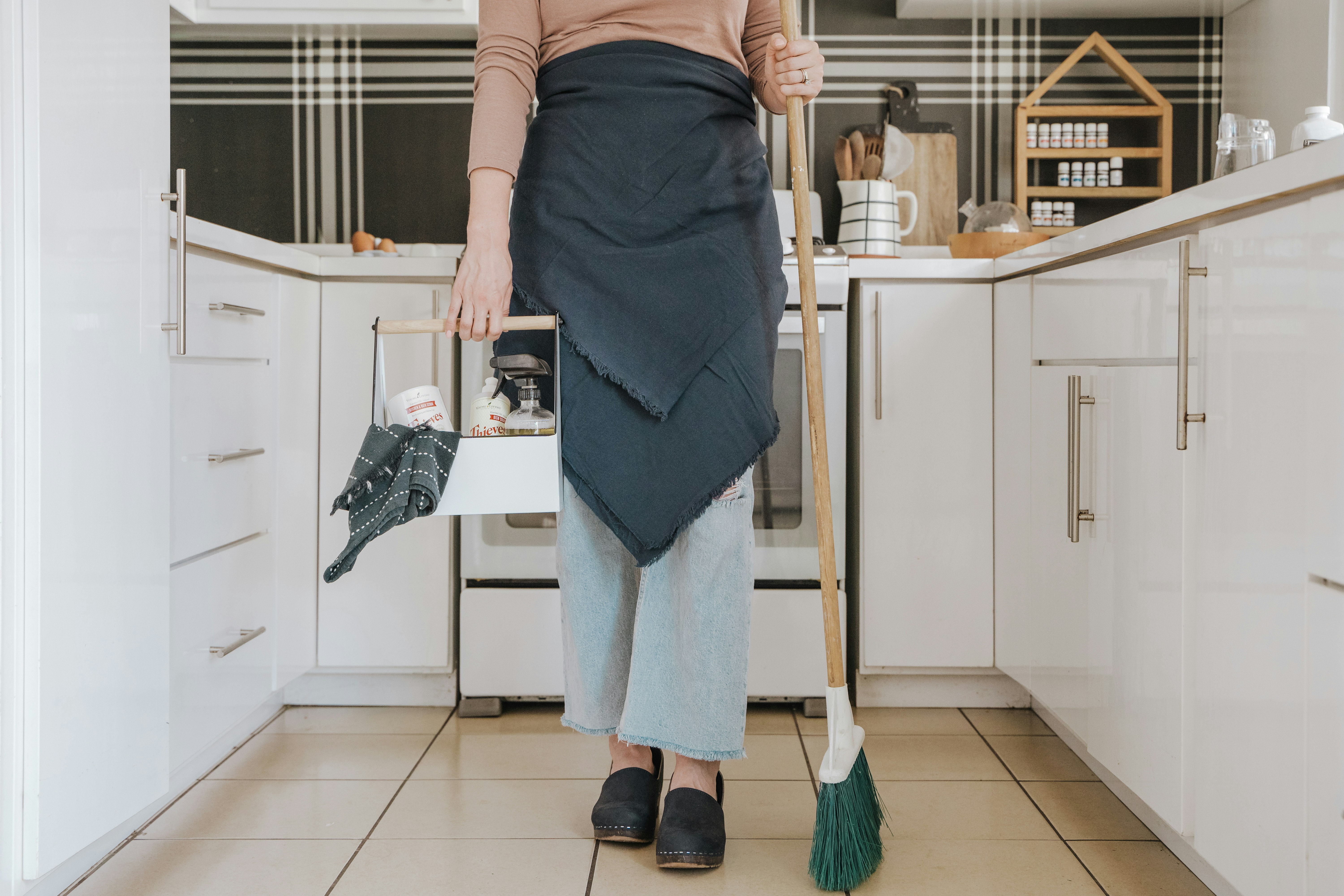 Pessoa em p&eacute; na cozinha, segurando uma caixa de produtos de limpeza e uma vassoura, pronta para limpar. Ela veste blusa bege, avental azul-marinho e cal&ccedil;a jeans clara, com sapatos pretos. A cozinha tem arm&aacute;rios brancos e azulejos quadriculados em preto e branco na parede