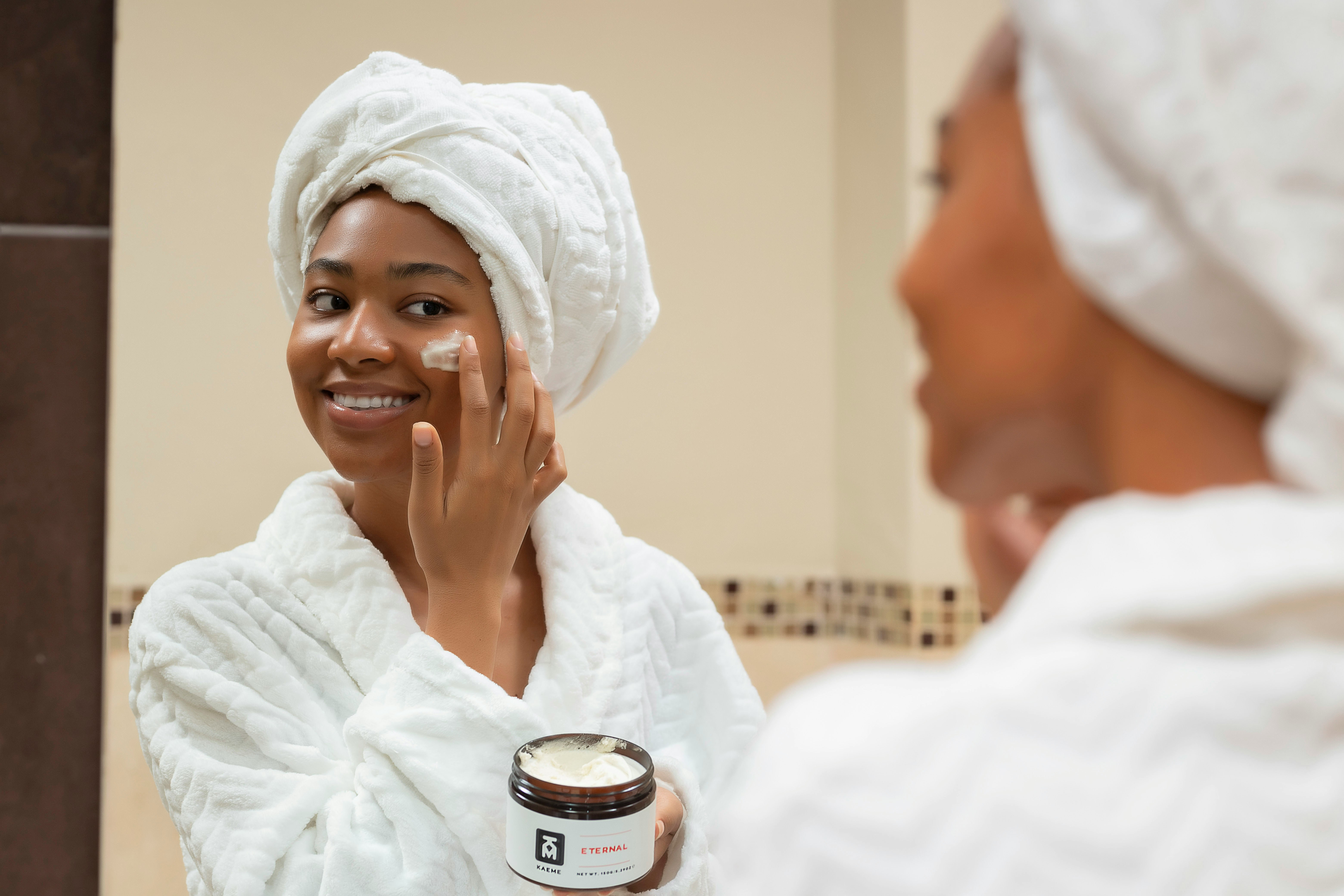 Mulher negra sorridente, com toalha branca na cabe&ccedil;a e roup&atilde;o, aplica creme facial no rosto enquanto se olha no espelho, segurando um pote de creme com a m&atilde;o esquerda