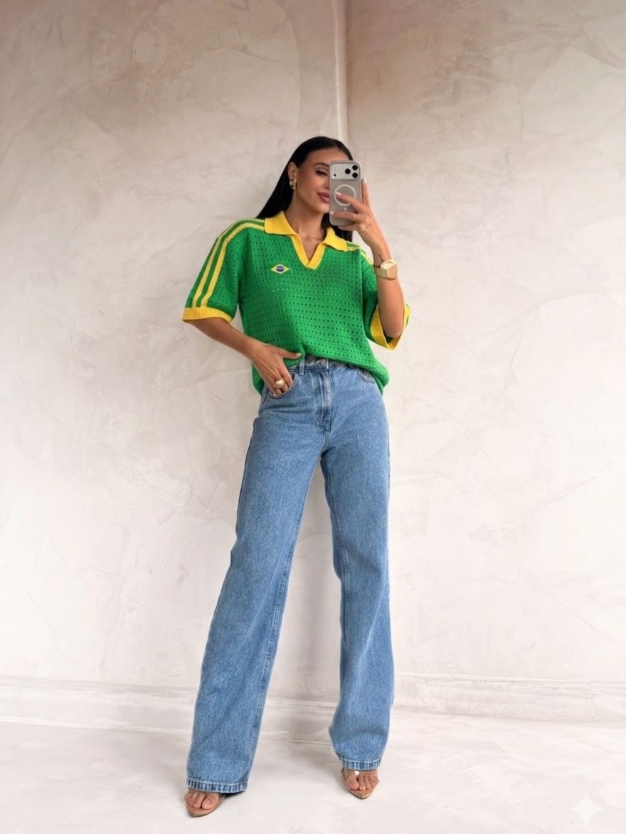 Mulher de cabelos escuros, usando blusa de malha verde com gola e listras amarelas, e um pequeno emblema da bandeira do Brasil. Ela veste cal&ccedil;a jeans clara de corte reto, sand&aacute;lias de salto baixo e segura um celular para tirar uma selfie, com a m&atilde;o esquerda no bolso. O fundo &eacute; uma parede clara com textura.