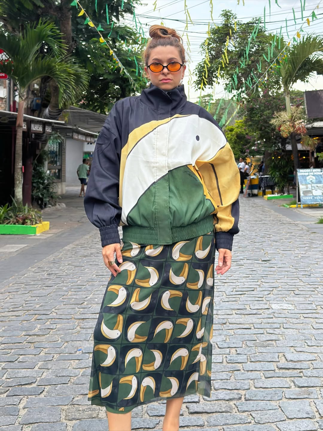 Mulher de cabelo preso em coque, &oacute;culos laranja, jaqueta bomber preta, amarela, branca e verde com estampa de tucano, e saia longa verde escura com tucanos estilizados, posando em rua de paralelep&iacute;pedos com palmeiras e bandeirinhas de festa junina ao fundo