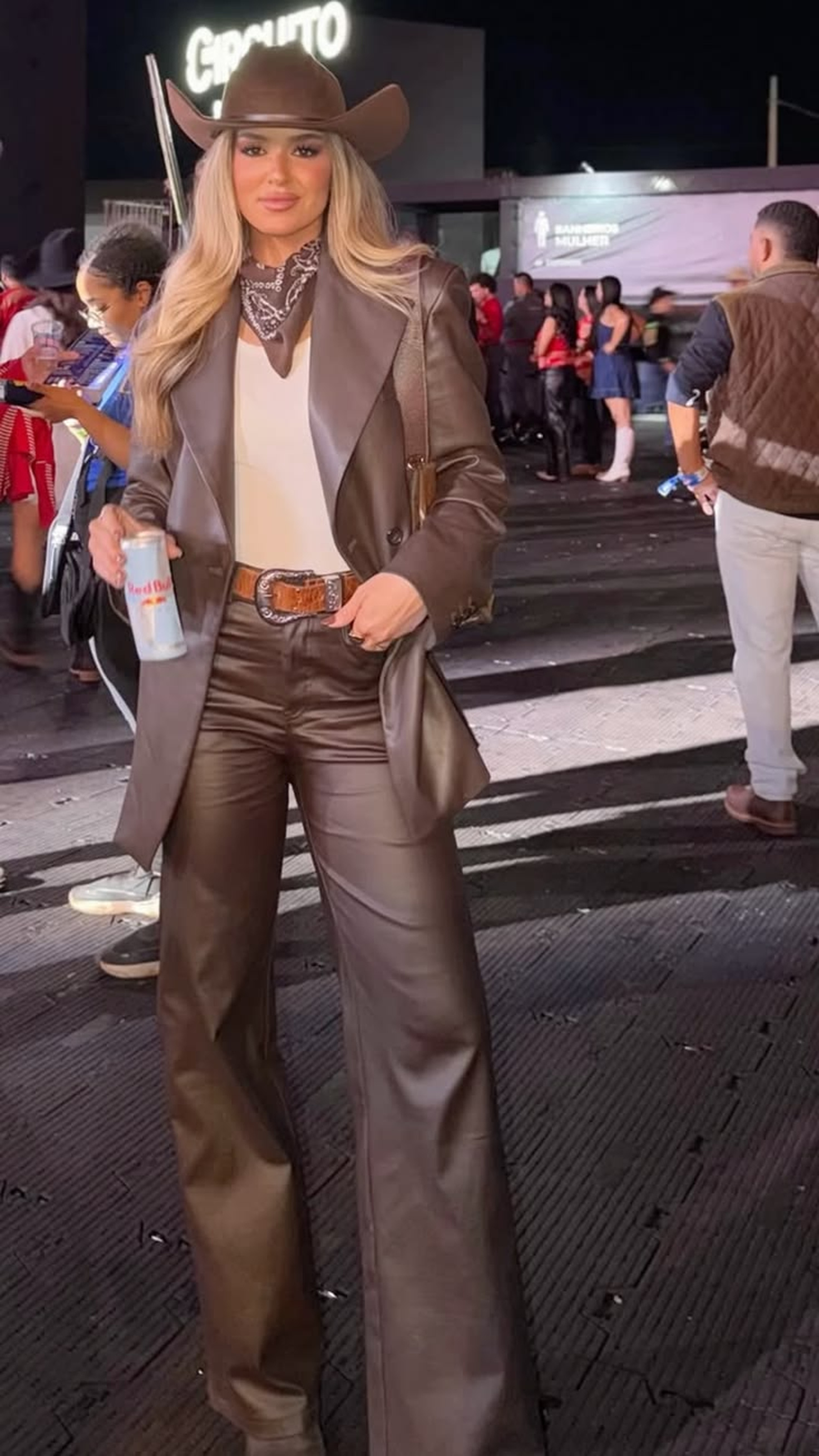 Mulher loira sorrindo, usando chap&eacute;u e bandana marrons, blazer e cal&ccedil;a de couro marrons, cinto com fivela grande, segurando uma lata de Red Bull em um evento noturno com outras pessoas e um letreiro CIRCUITO ao fundo