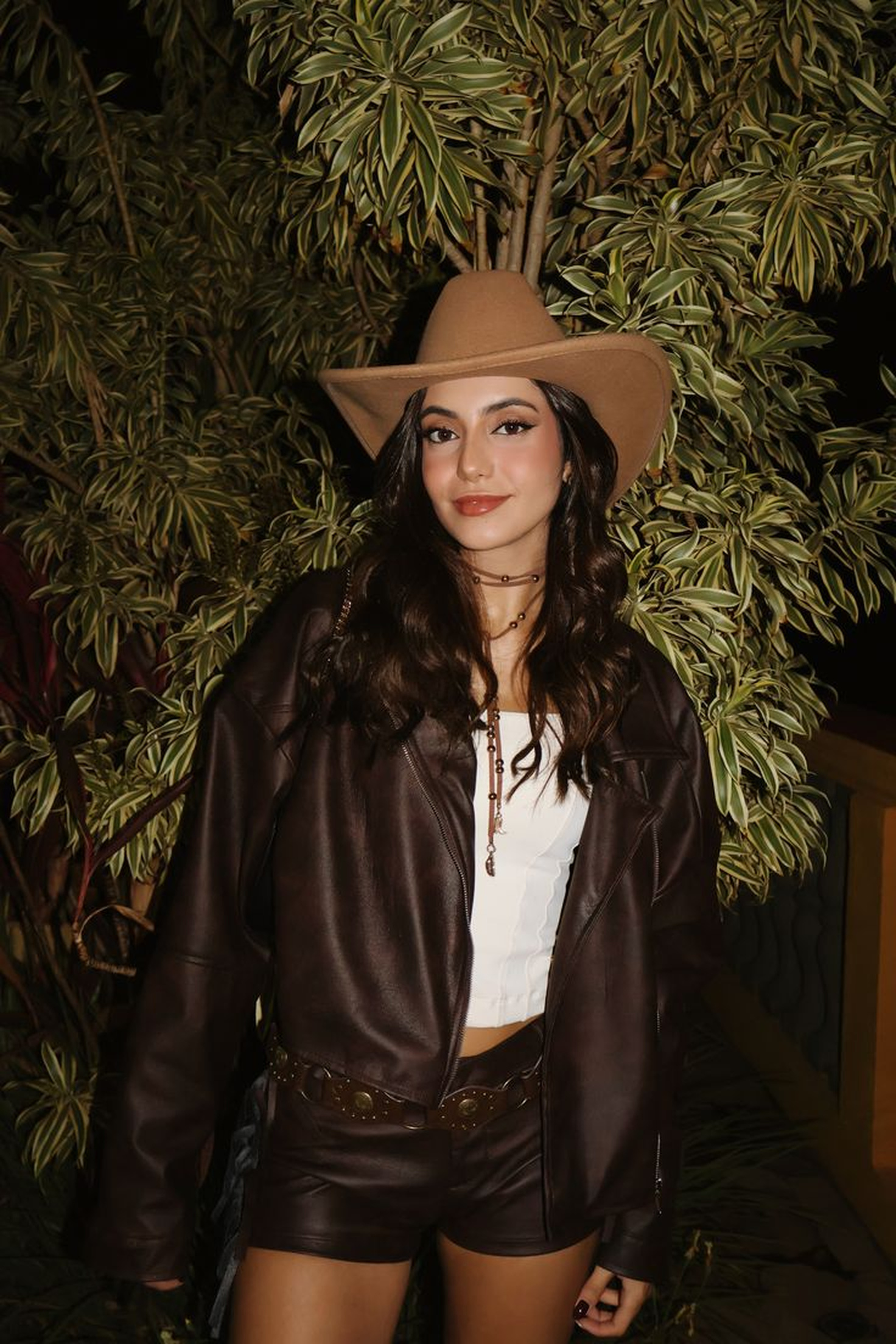 Mulher jovem com chap&eacute;u caub&oacute;i marrom, jaqueta de couro marrom, top branco e shorts de couro, sorrindo em frente a folhagens verdes e escuras