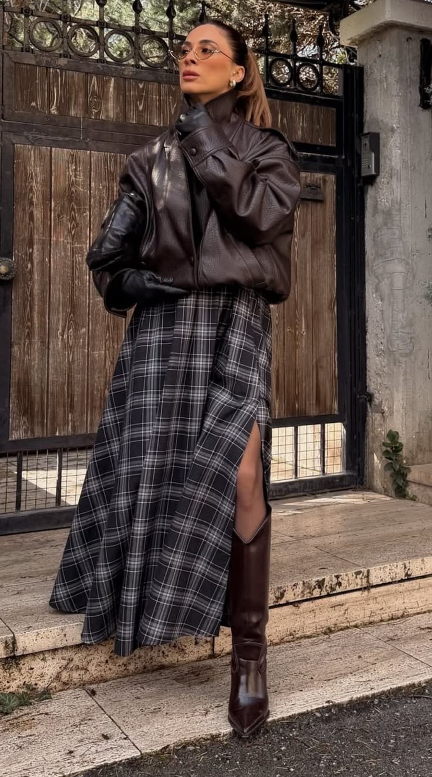 Mulher de &oacute;culos redondos, jaqueta de couro marrom, luvas pretas e saia xadrez preta e branca com fenda, posando em frente a um port&atilde;o de madeira e uma parede de concreto, com botas de cano alto marrons vis&iacute;veis