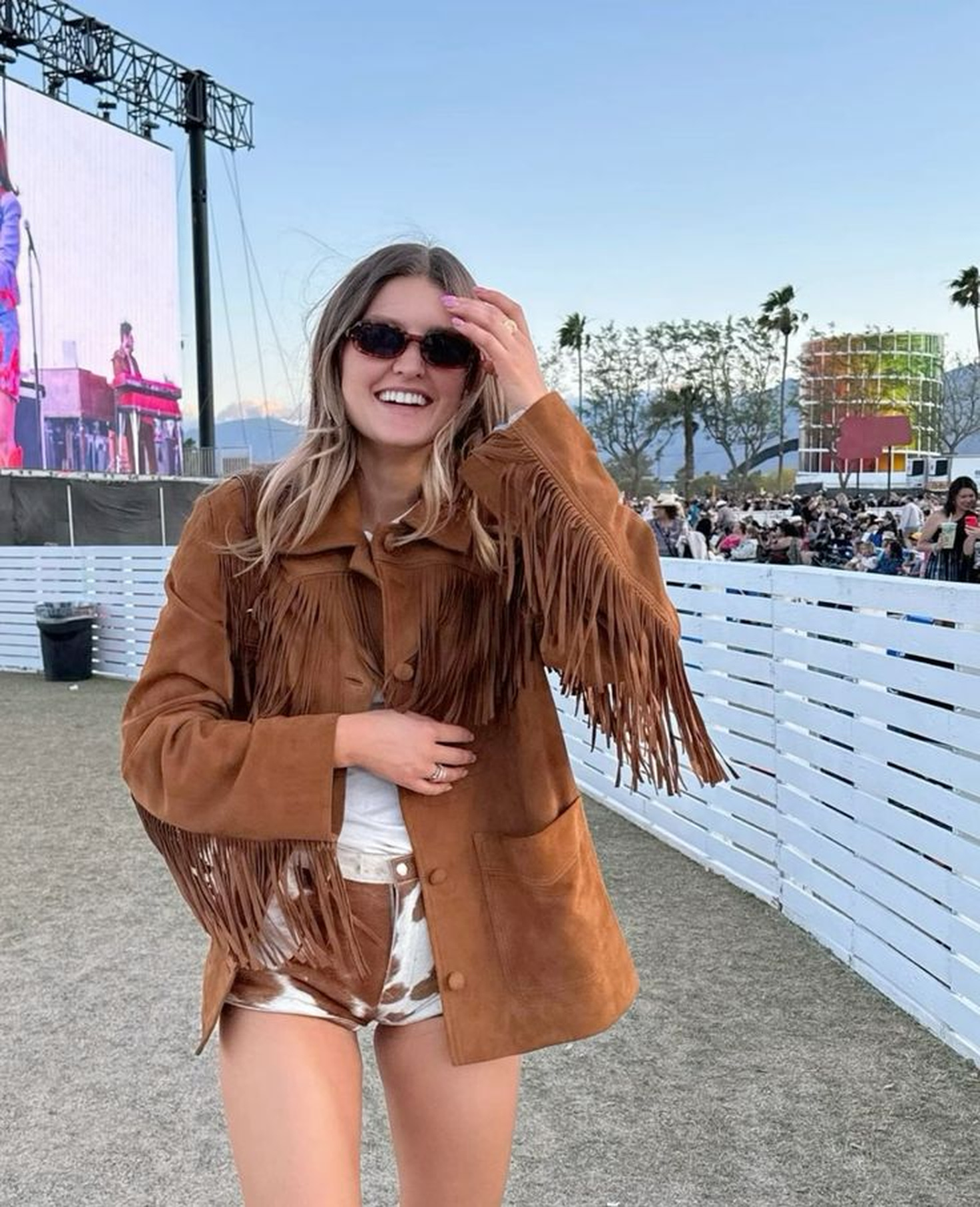 Mulher loira sorrindo, usando &oacute;culos escuros, jaqueta de camur&ccedil;a marrom com franjas e shorts de estampa de vaca, em festival ao ar livre com palco e p&uacute;blico ao fundo