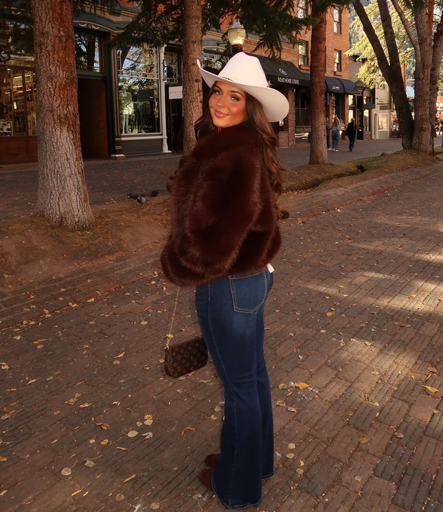 Mulher sorridente, com chap&eacute;u de cowboy branco e casaco marrom de pelos, posa em rua de paralelep&iacute;pedos com &aacute;rvores e pr&eacute;dios de tijolos ao fundo