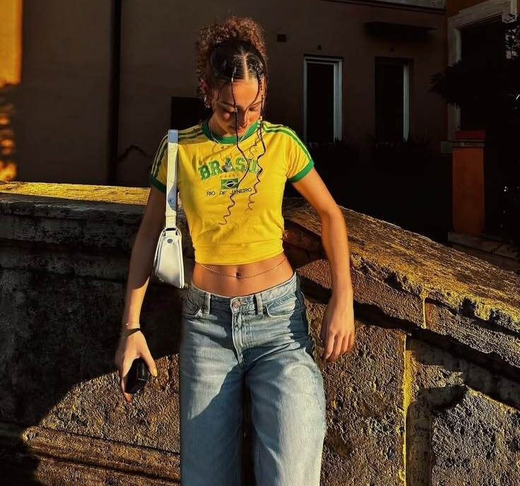 Jovem mulher com cabelo cacheado preso, vestindo camiseta amarela do Brasil e cal&ccedil;a jeans larga, apoiada em muro de pedra sob luz dourada do p&ocirc;r do sol