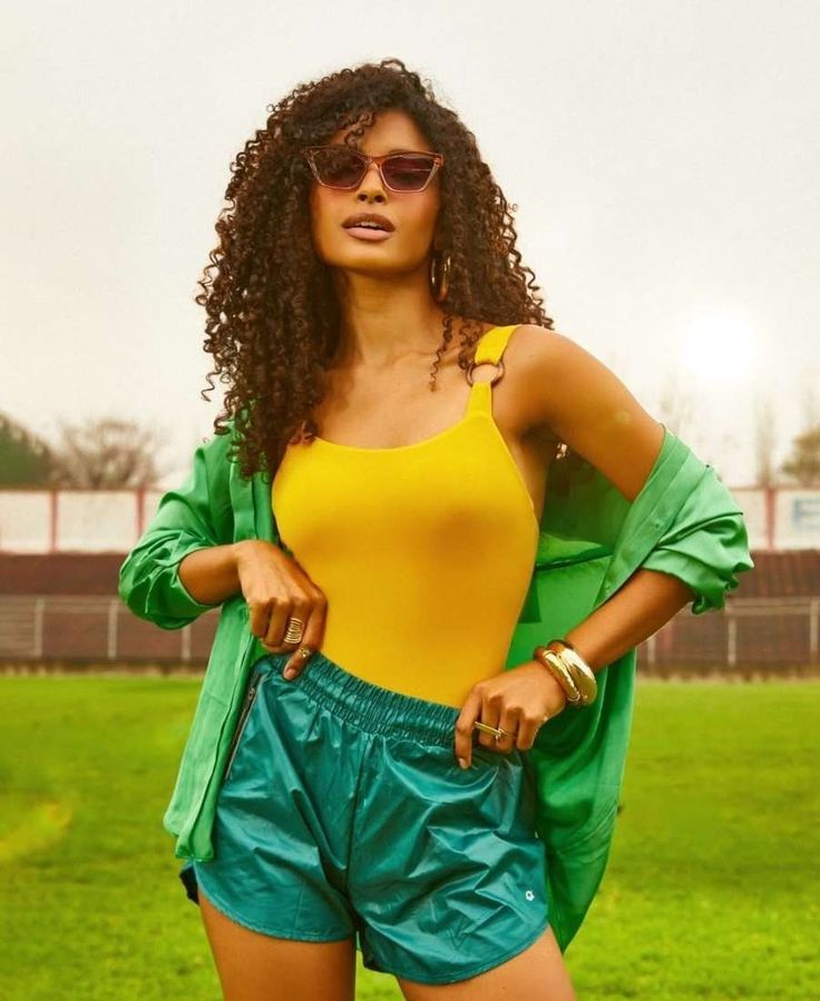 Mulher de pele morena e cabelos cacheados longos, usando &oacute;culos de sol, blusa amarela, shorts verde-&aacute;gua e camisa verde aberta, posando em um campo gramado