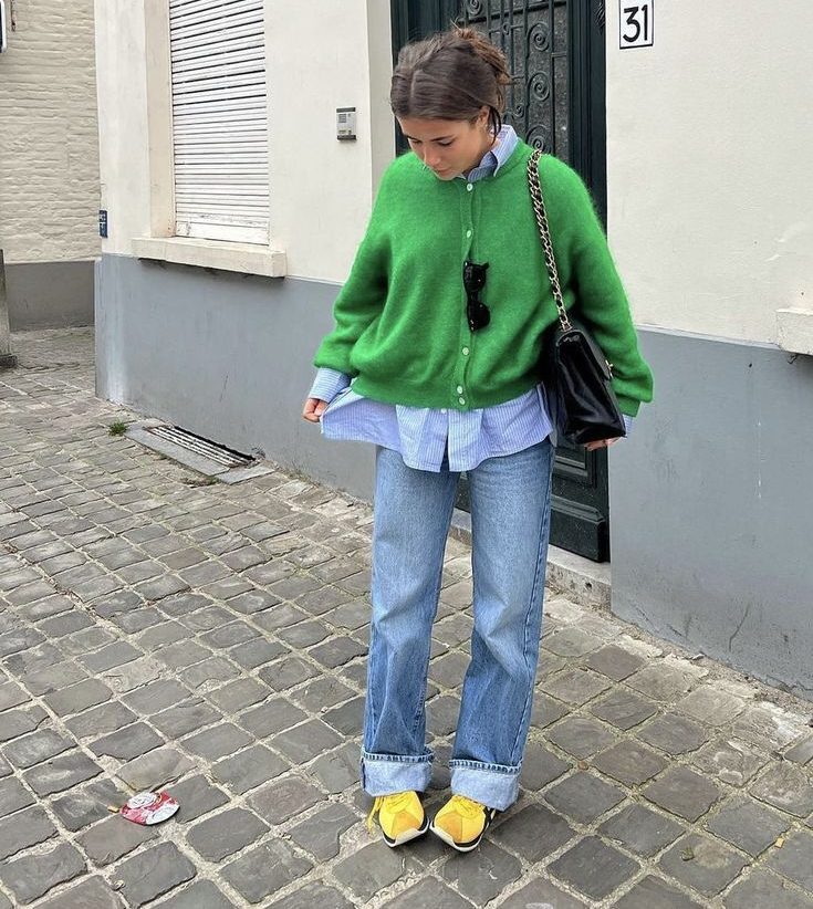 Mulher jovem com cabelo preso, usando cardig&atilde; verde, camisa listrada azul e branca, cal&ccedil;a jeans larga com barra dobrada, t&ecirc;nis amarelos e bolsa preta de ombro, em cal&ccedil;ada de paralelep&iacute;pedos com fachada de pr&eacute;dio branco e porta verde escura ao fundo